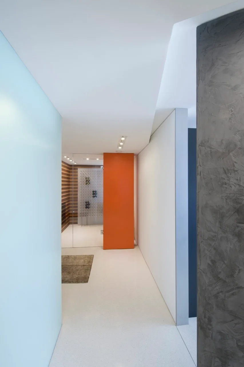 A minimalist white hallway with a textured grey plaster wall leads to a glass-enclosed wine room featuring a bold orange partition panel.