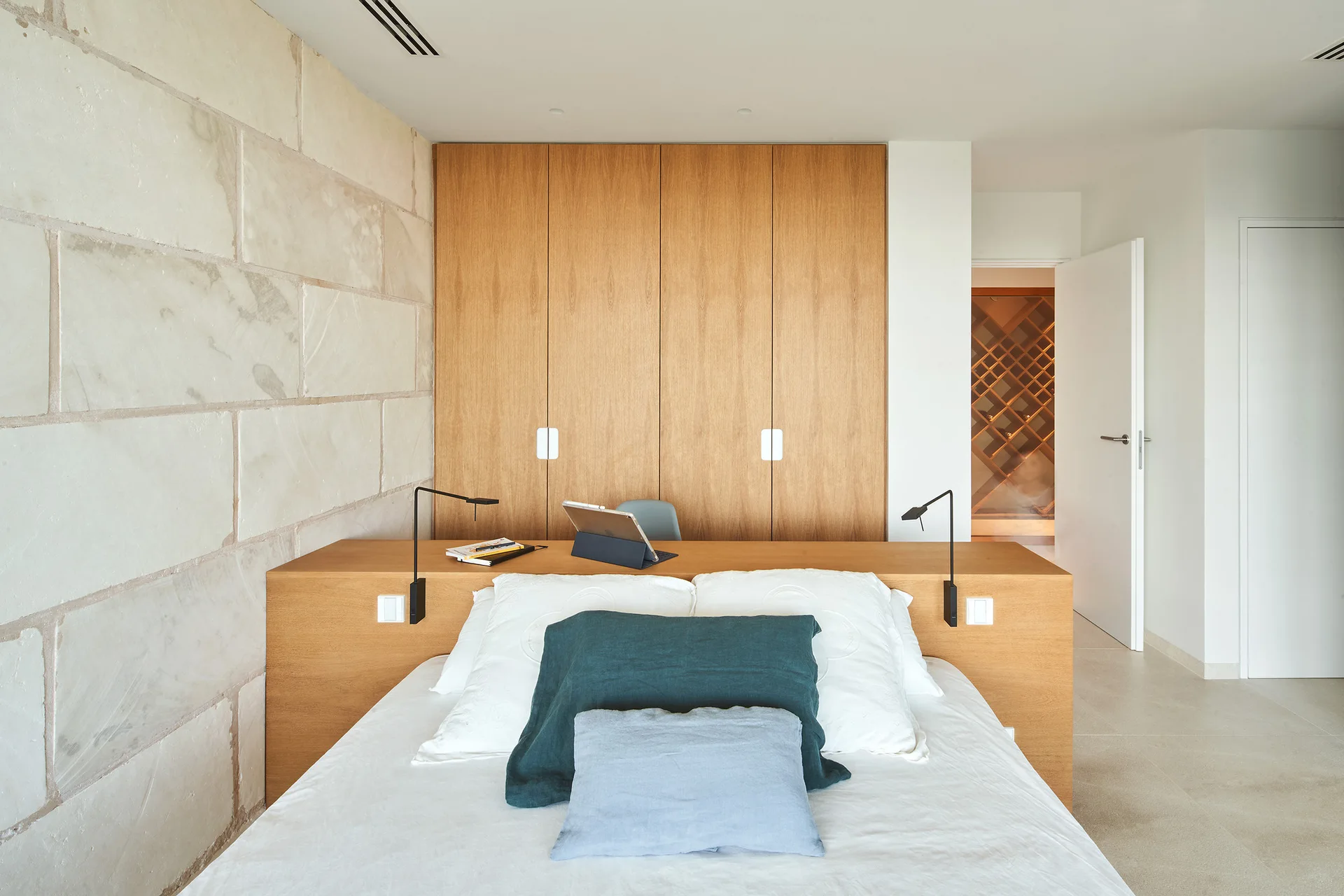 A minimalist bedroom featuring an integrated wooden headboard desk, a natural stone block accent wall, and a light wood wardrobe.