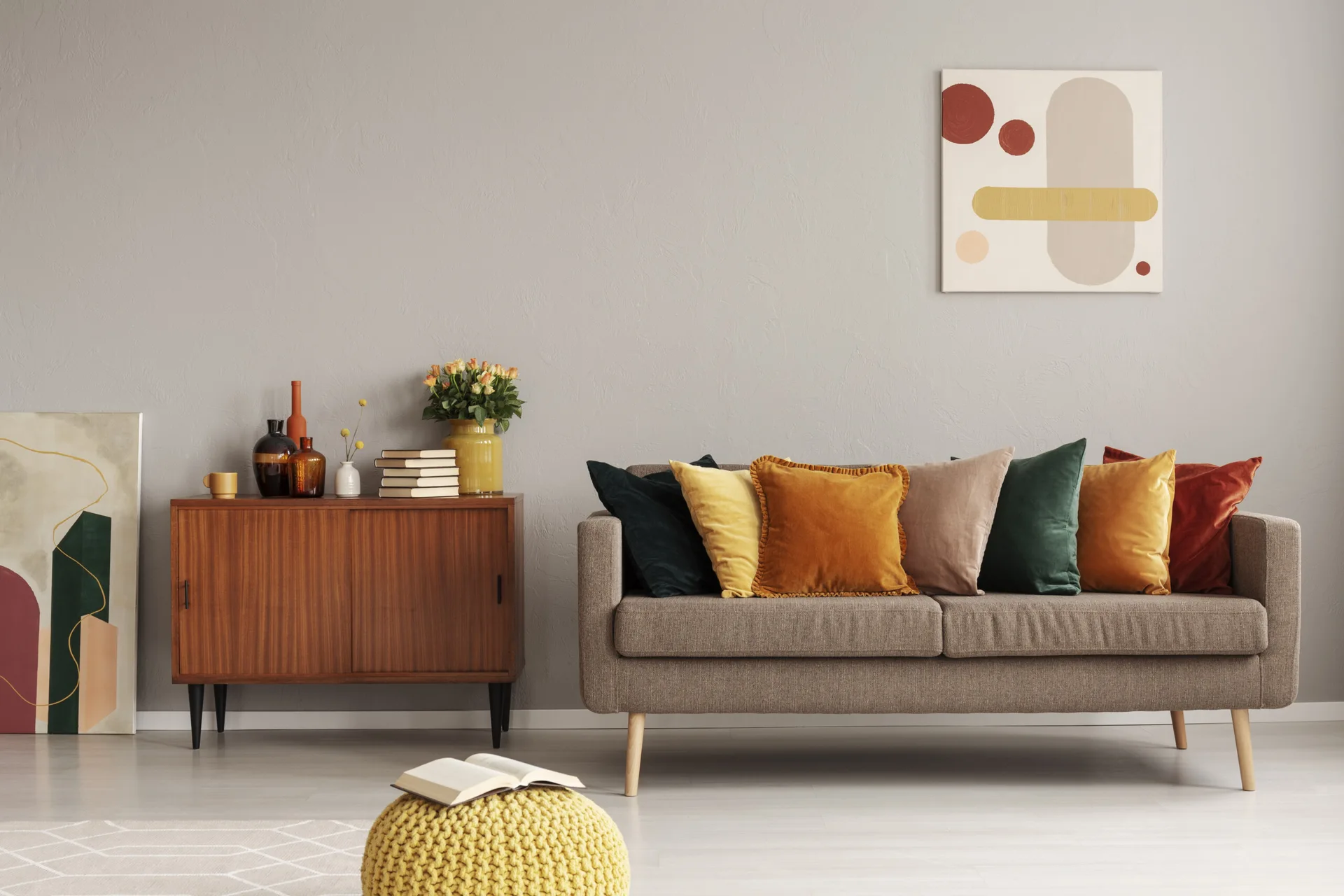 A grey sofa with colorful velvet pillows stands next to a wooden sideboard and a yellow knitted ottoman in a modern living room.