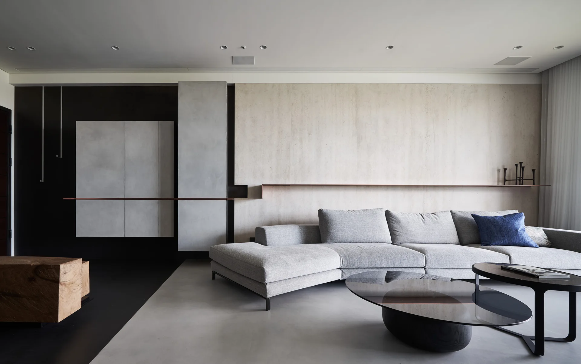 A grey sectional sofa sits before a textured beige paneled wall featuring a thin copper-toned ledge and a dark glass coffee table.