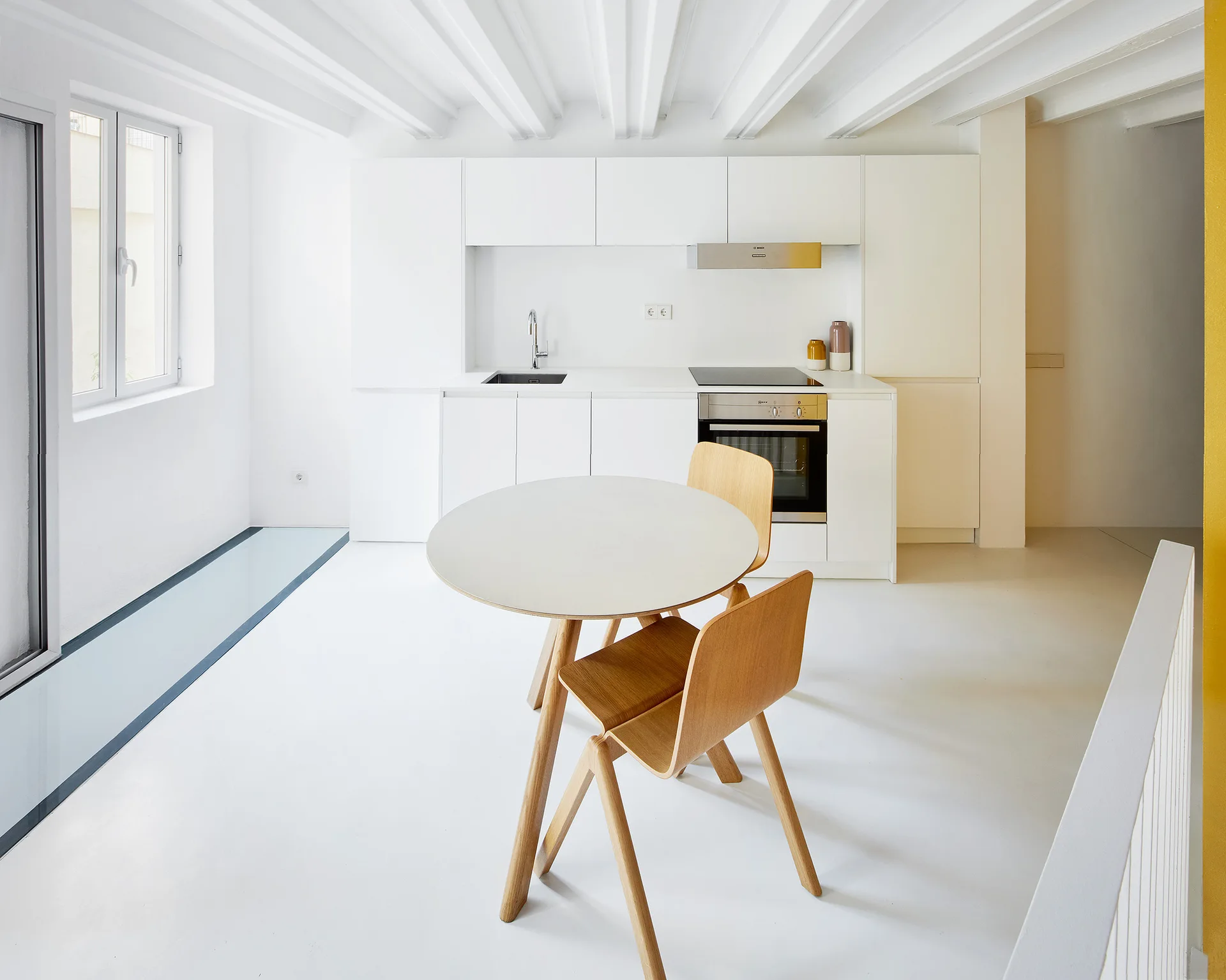 A minimalist white kitchen and dining area features a round table with wood chairs, white flat-front cabinets, and exposed ceiling beams.