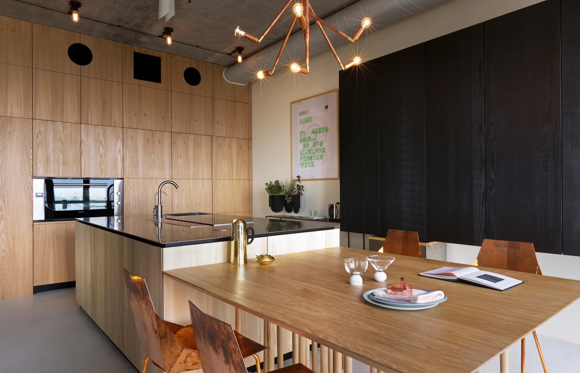 A modern kitchen with light oak and black wood cabinets, an island with an integrated wood dining table, copper chairs, and an exposed concrete ceiling.