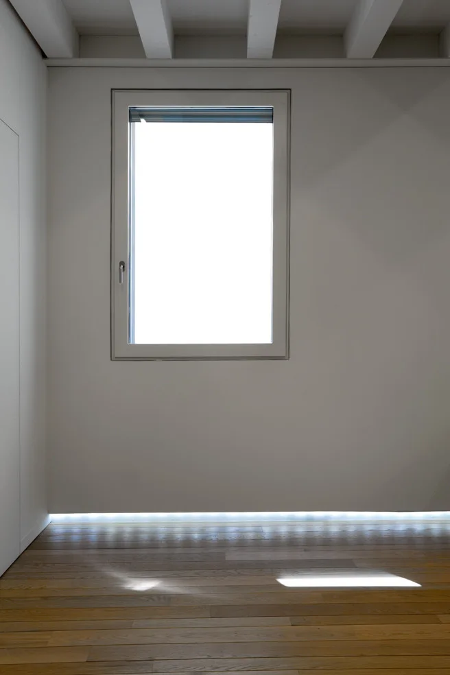 Minimalist white wall with a rectangular window and a glowing LED light strip recessed along the baseboard of the wood plank floor.