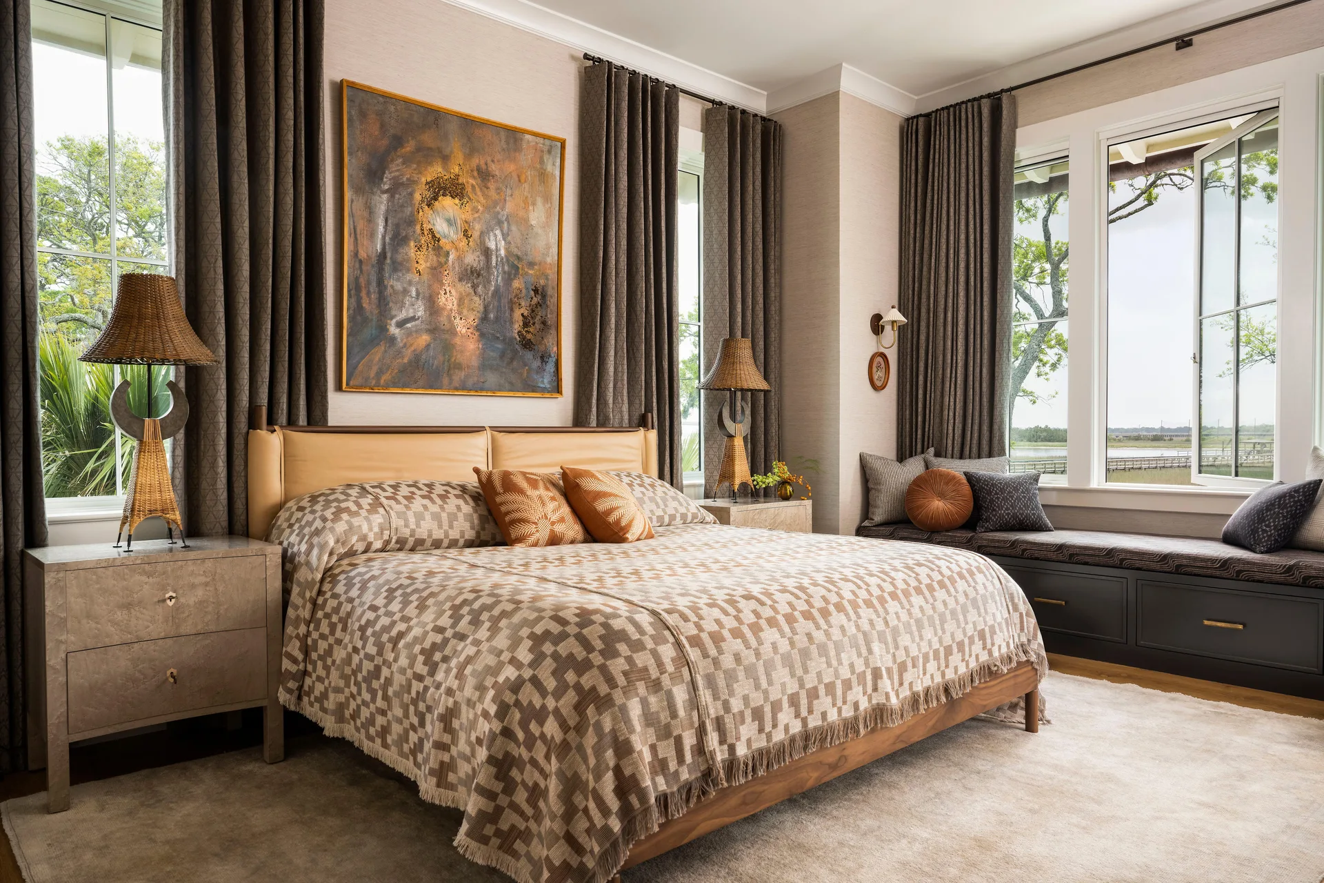 A neutral bedroom features a bed with geometric brown bedding, paired with textured wallpaper, woven table lamps, and a built-in window bench.