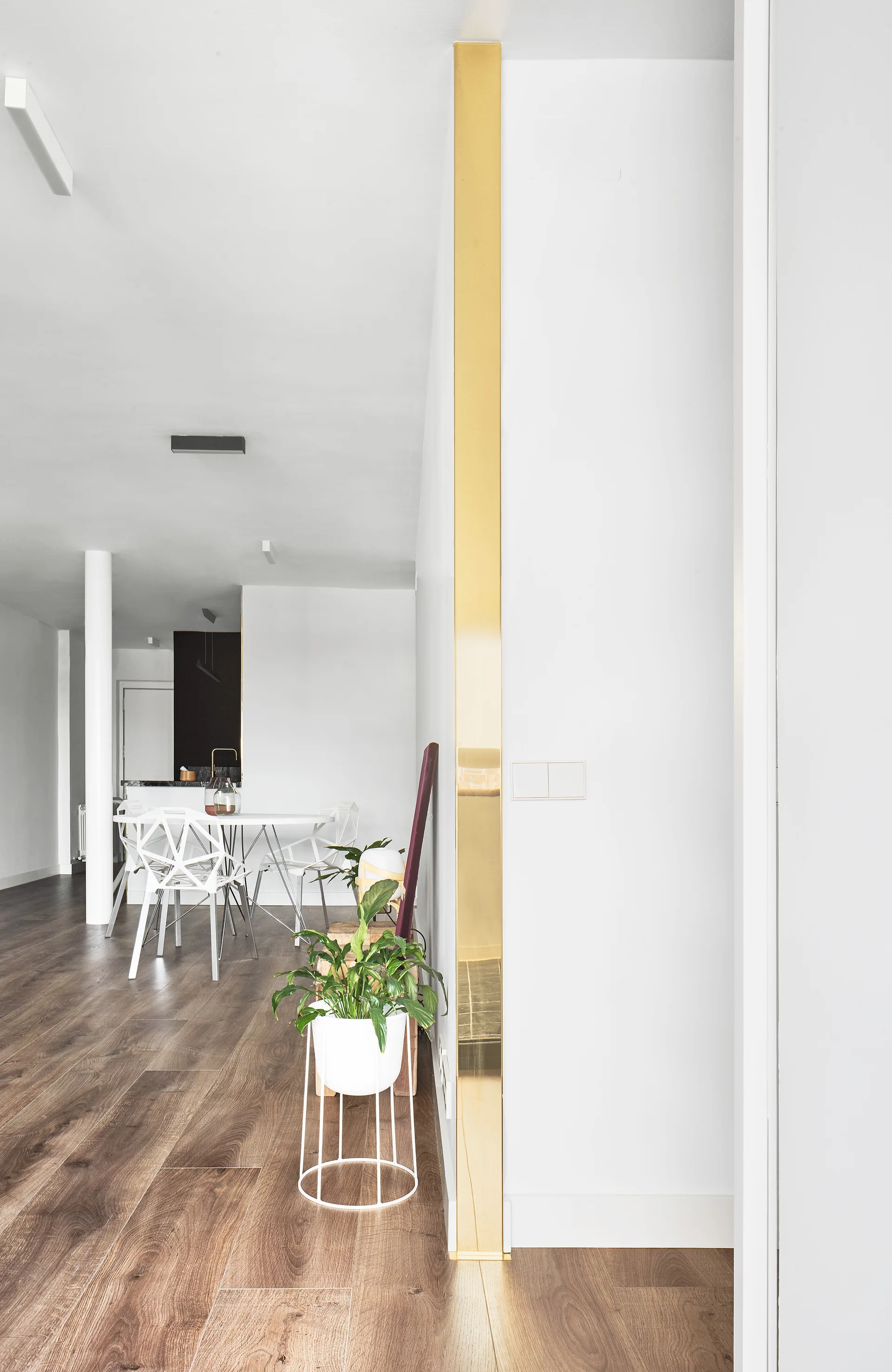A vertical gold metal trim accents a white wall corner next to a minimalist dining set with geometric white chairs on dark wood floors.