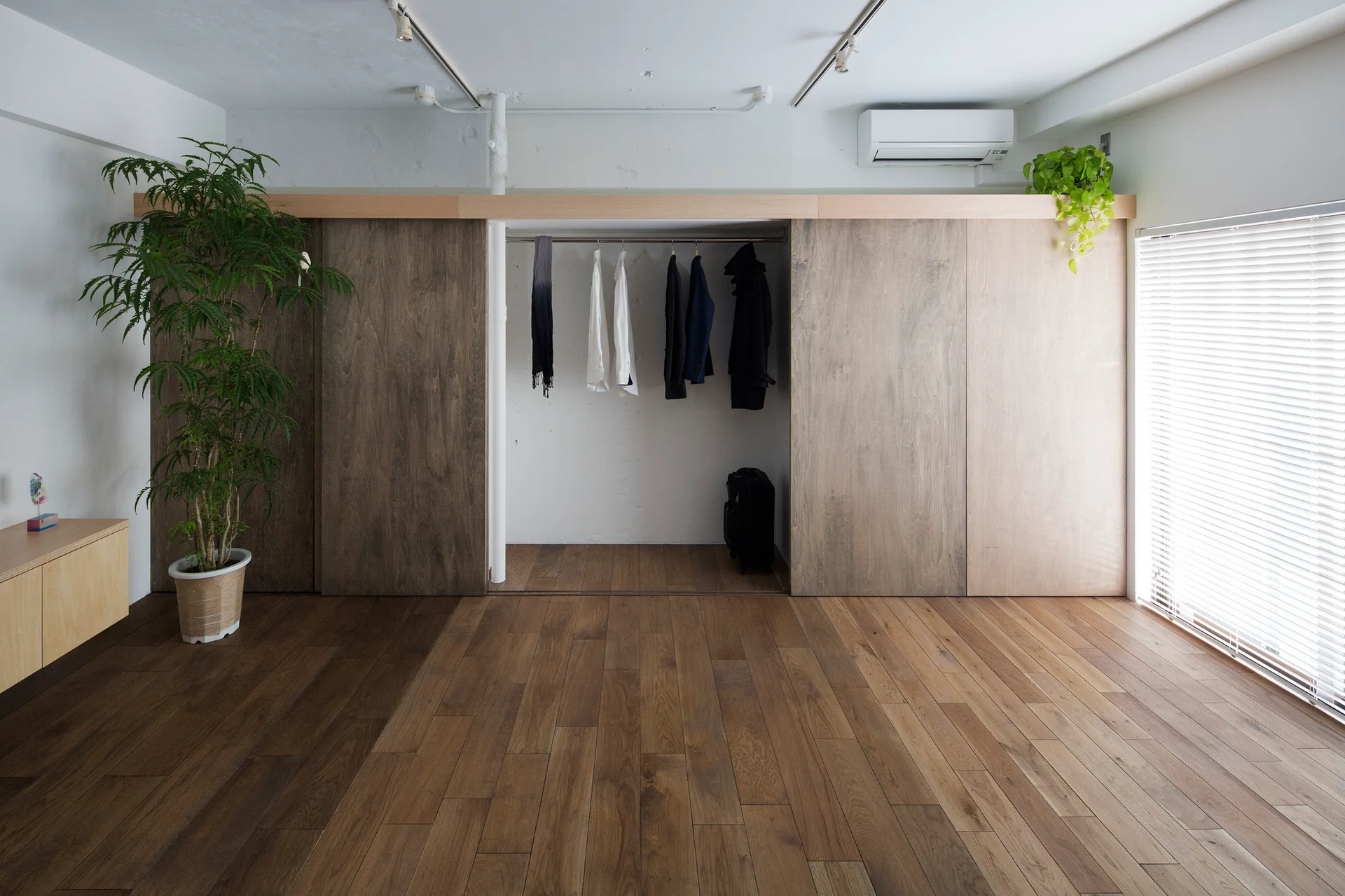 A minimalist room featuring a large wood-paneled wardrobe with open center hanging space and wide plank wood floors next to a window with blinds.