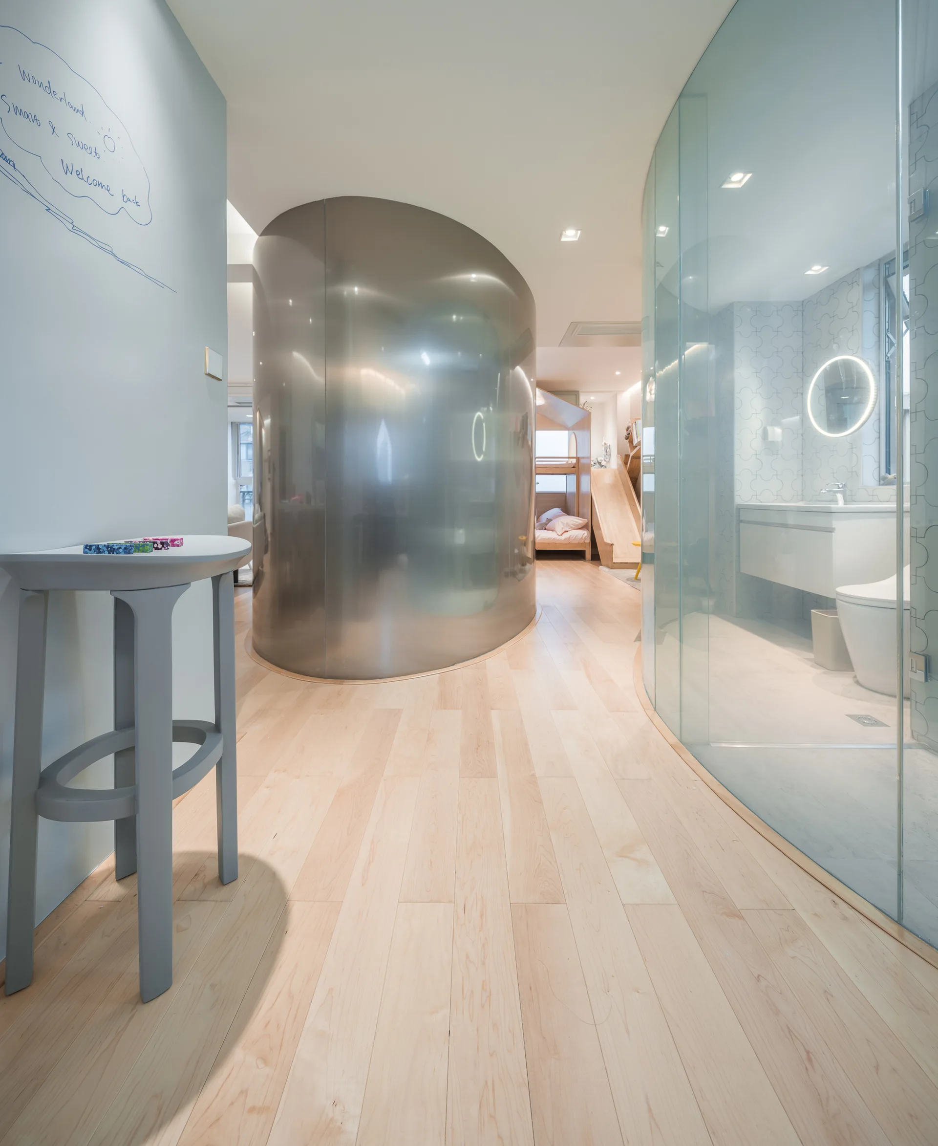 A modern hallway featuring a curved brushed metal partition wall, light wood flooring, and a glass-enclosed bathroom.