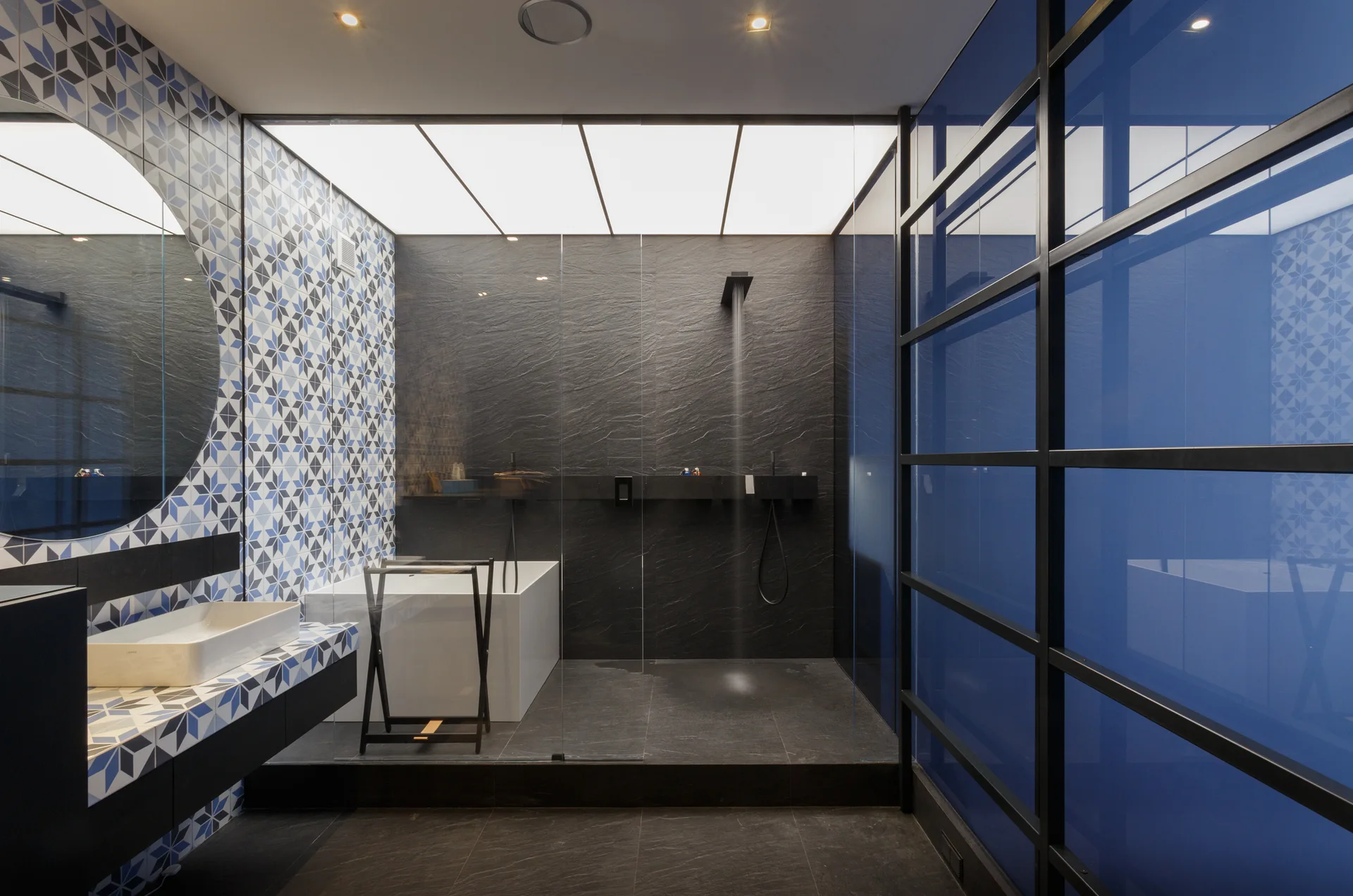 A modern bathroom features a dark slate walk-in shower, a blue-tinted glass grid partition, and a geometric tiled vanity wall with a round mirror.