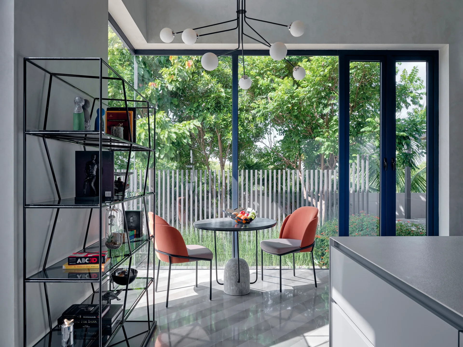 A modern dining corner features a black round table and two terracotta chairs set against a large window overlooking a lush garden.