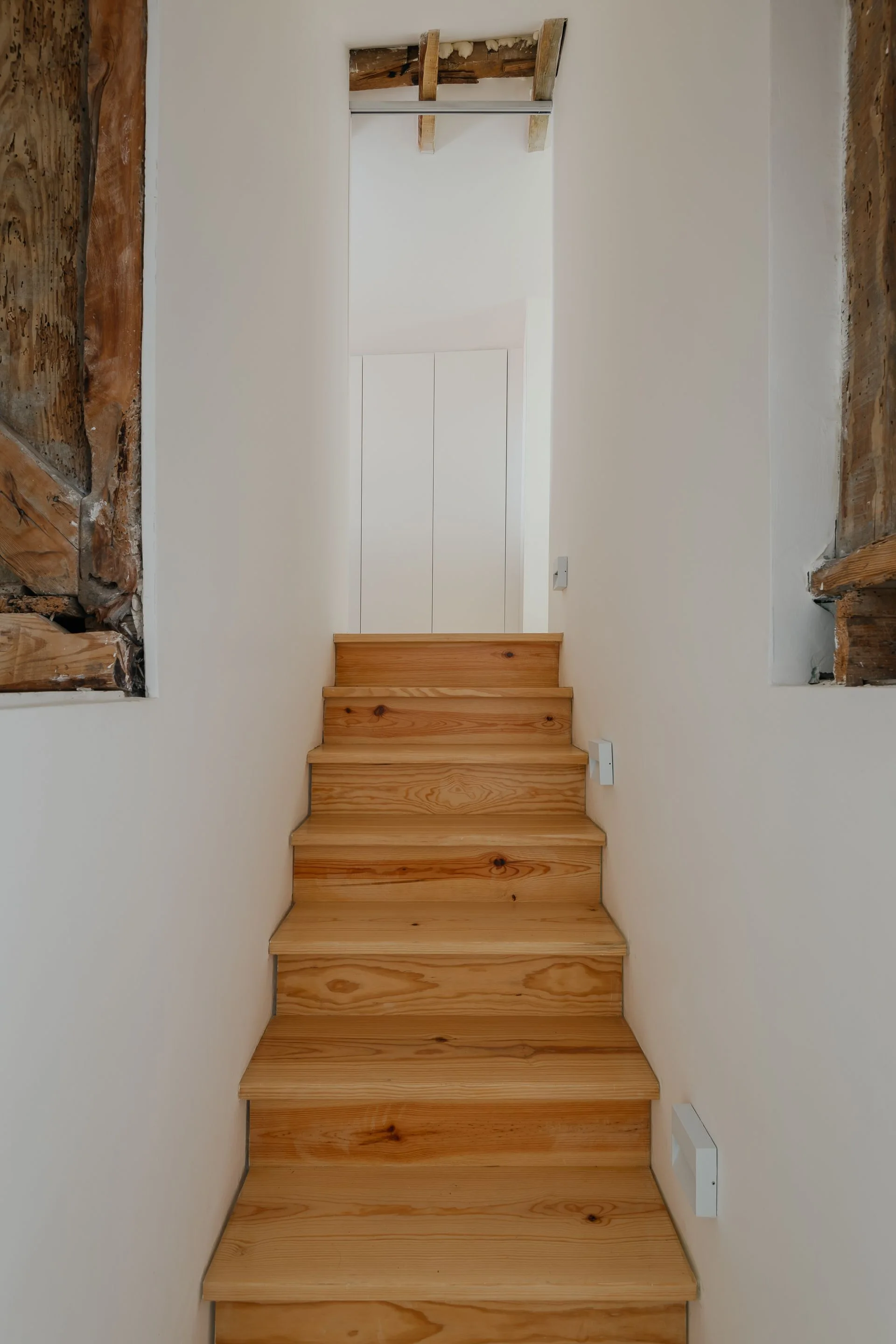 A narrow wood staircase ascends between smooth white walls that feature cutouts revealing rough rustic timber framing.