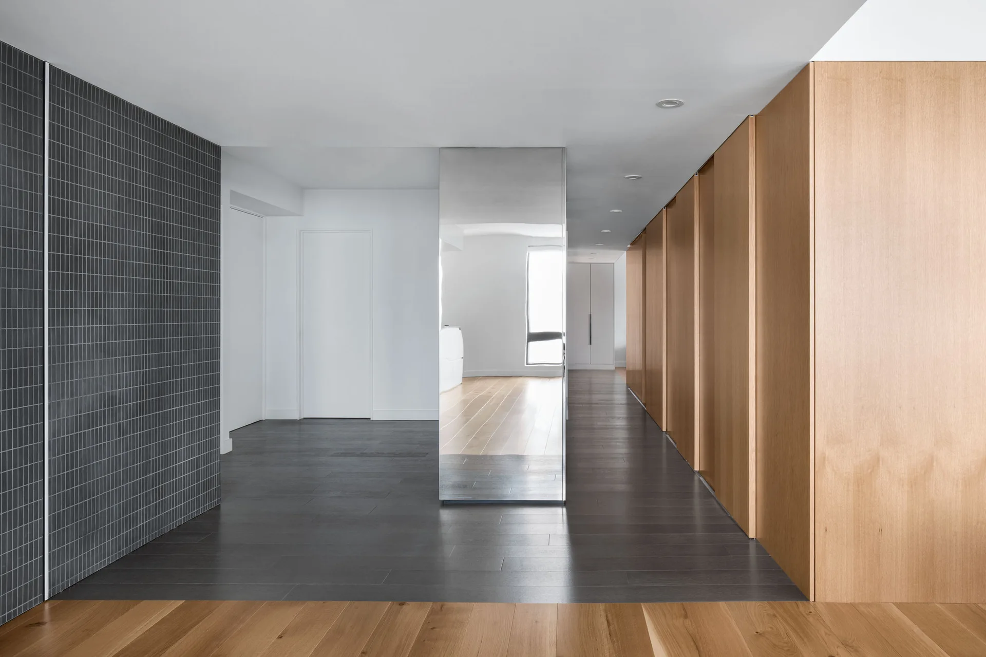 A modern hallway features natural wood panel closets, a dark grid-tiled wall, dark flooring, and a central floor-to-ceiling mirrored column.