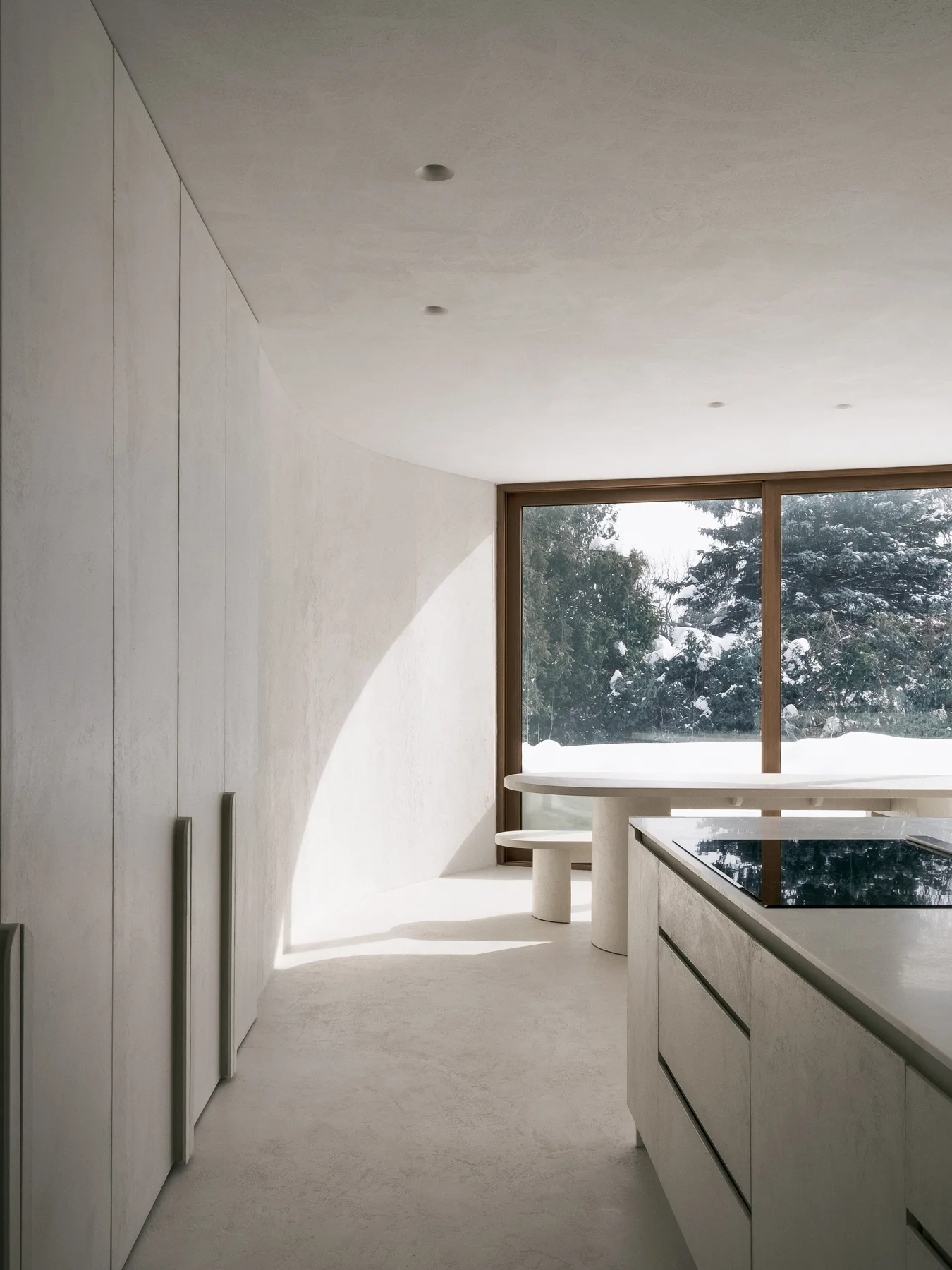 A minimalist kitchen features a curved plaster wall, matching greige cabinetry, and a large wood-framed window overlooking a snowy winter forest.
