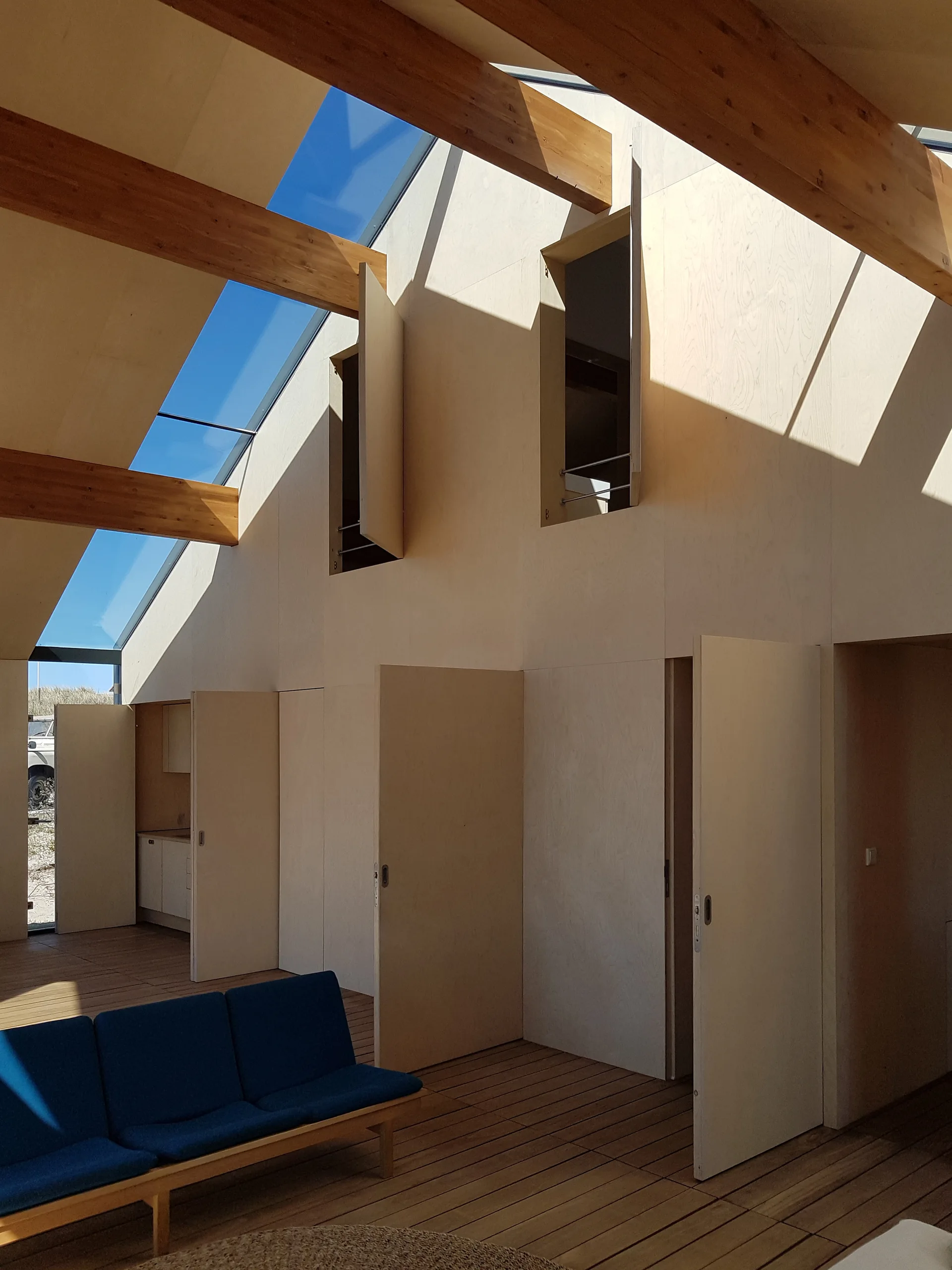 Minimalist living space featuring light plywood walls with integrated doors, exposed wood ceiling beams, a blue sofa, and large angled skylights.