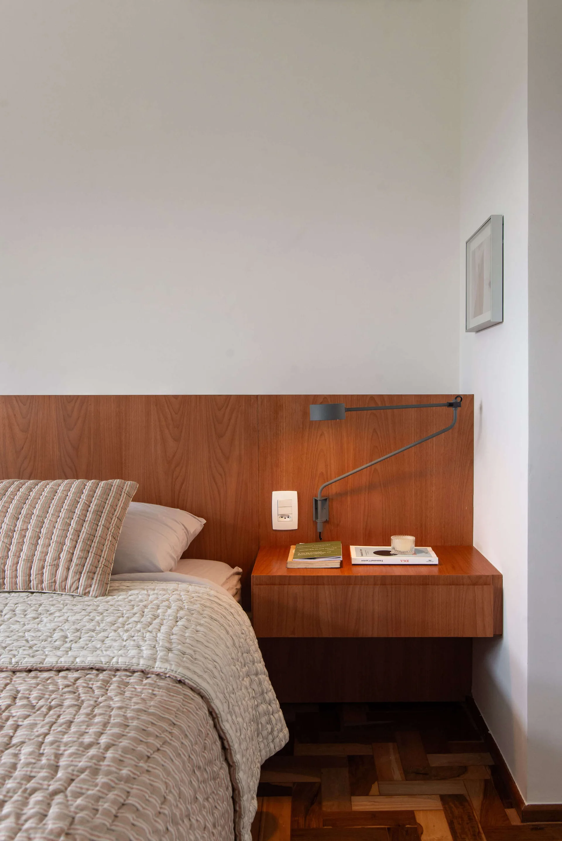 A bedroom corner featuring a wide wood panel headboard, a matching floating nightstand, a grey articulated wall lamp, and quilted bedding.