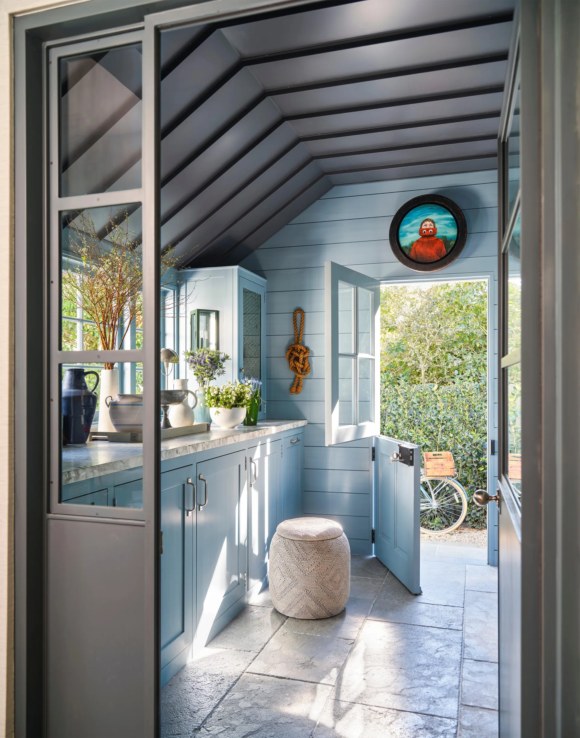 A blue-painted mudroom features light blue plank walls, grey stone flooring, a marble-topped counter, and an open Dutch door looking out to a garden.