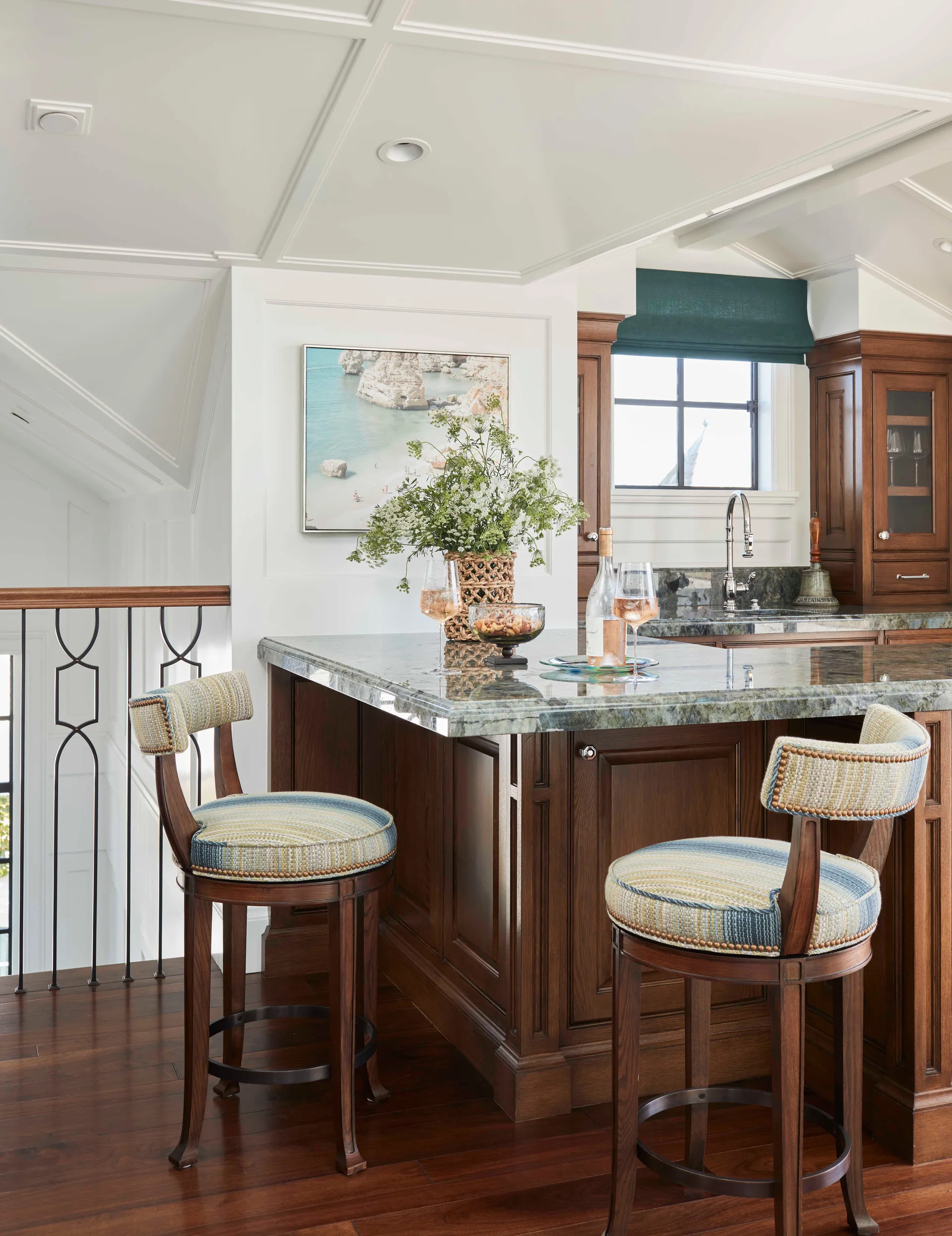 A dark wood kitchen island with a stone countertop features two striped upholstered bar stools next to a staircase with iron railings.