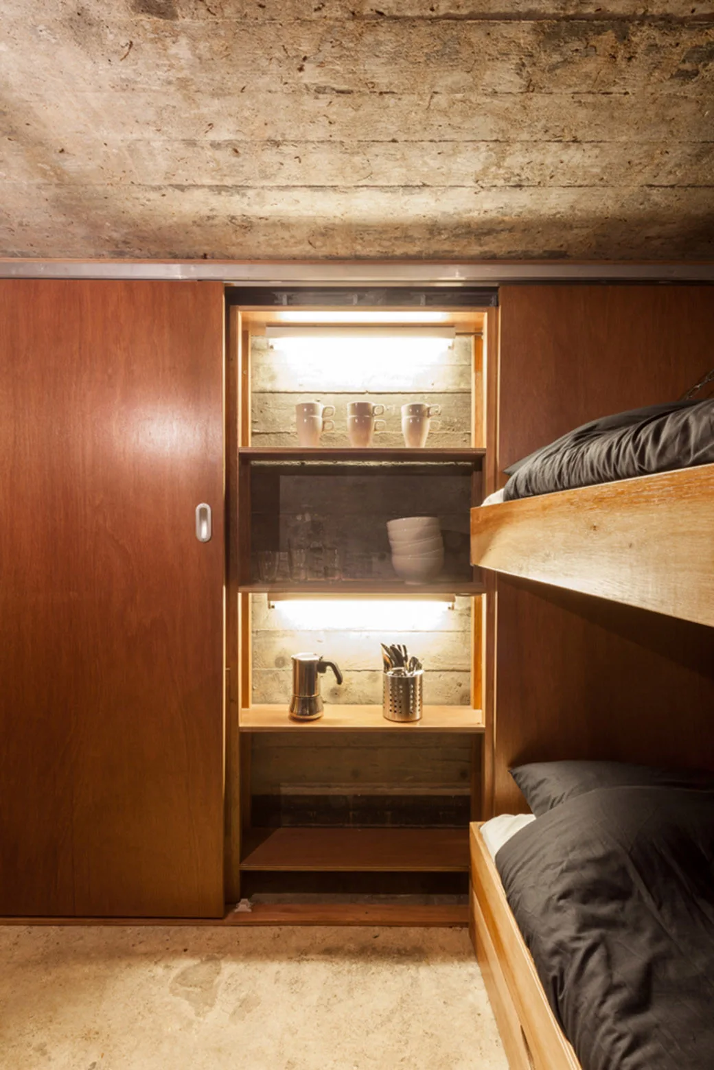 A compact bedroom featuring wooden bunk beds and a built-in wooden closet with a sliding door open to reveal lighted shelves against a concrete wall.