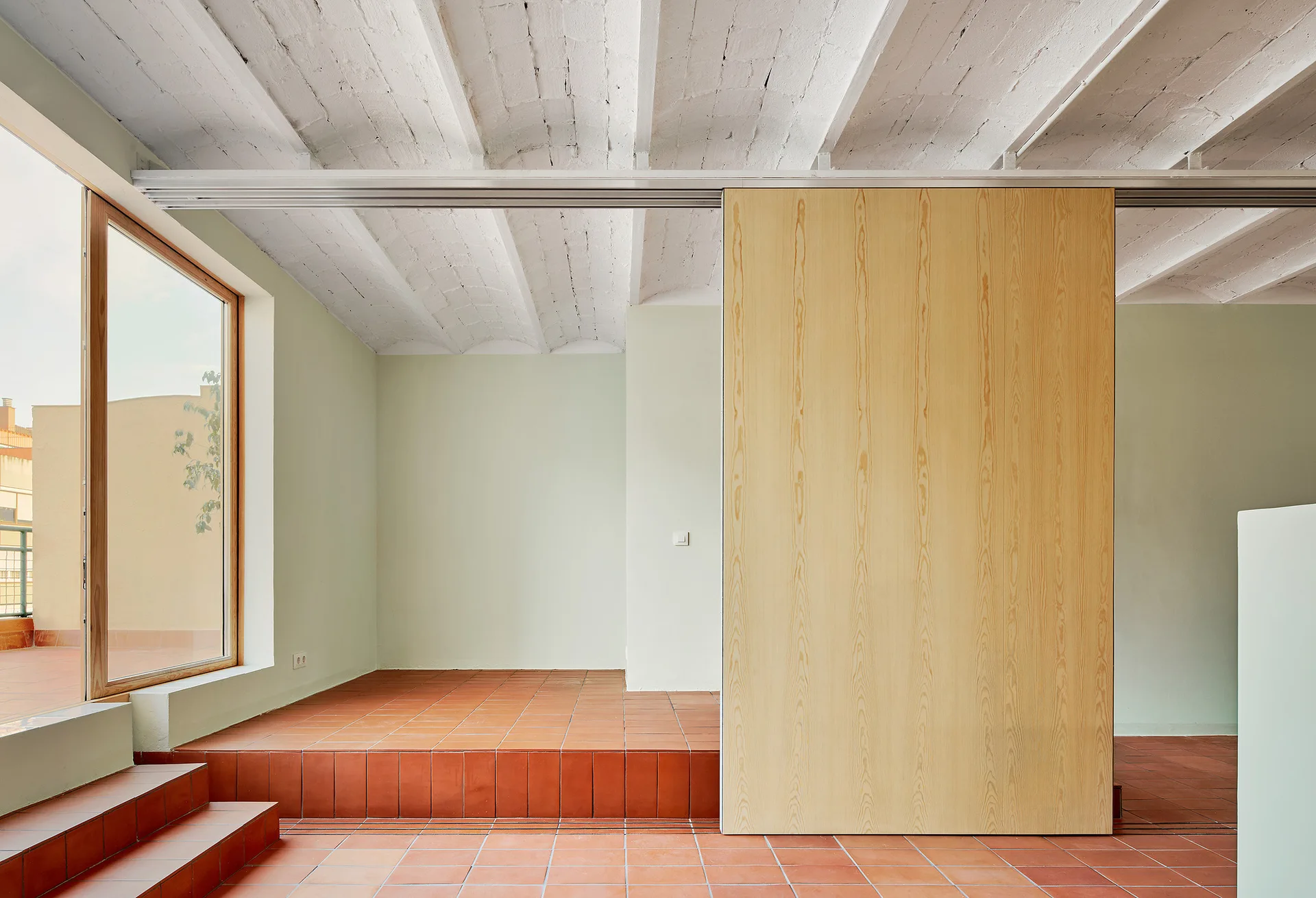 A minimalist room features pale green walls, a terracotta tiled floor with steps, a vaulted white ceiling, and a large wooden sliding partition.