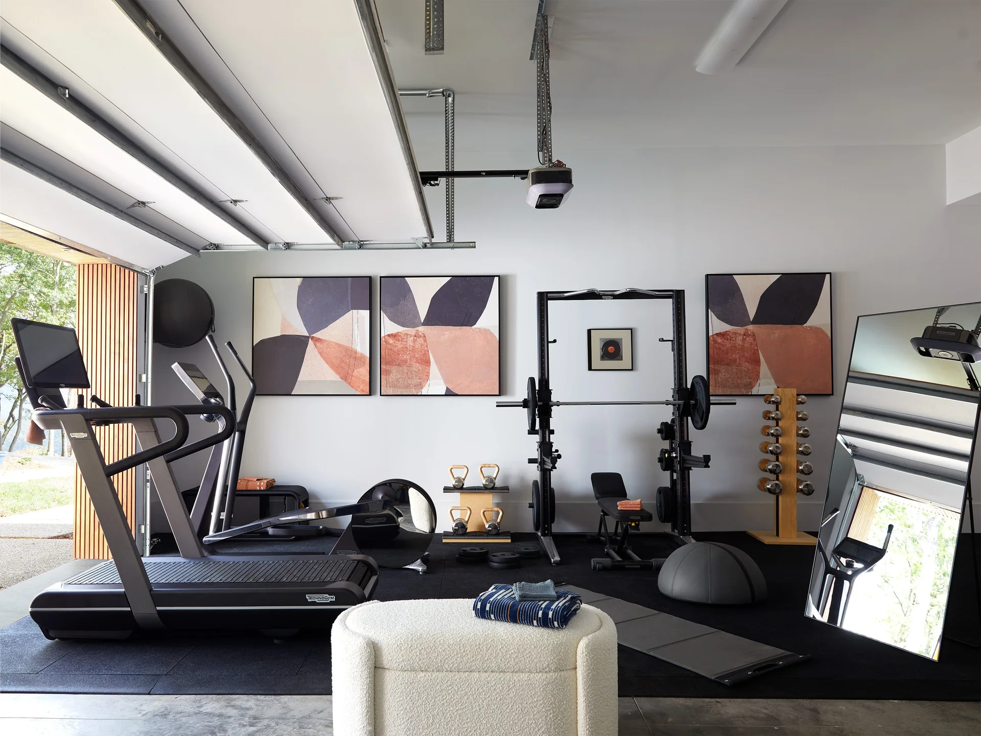 A modern home gym in a garage featuring black rubber flooring, weight training equipment, large abstract wall art, and an open garage door.