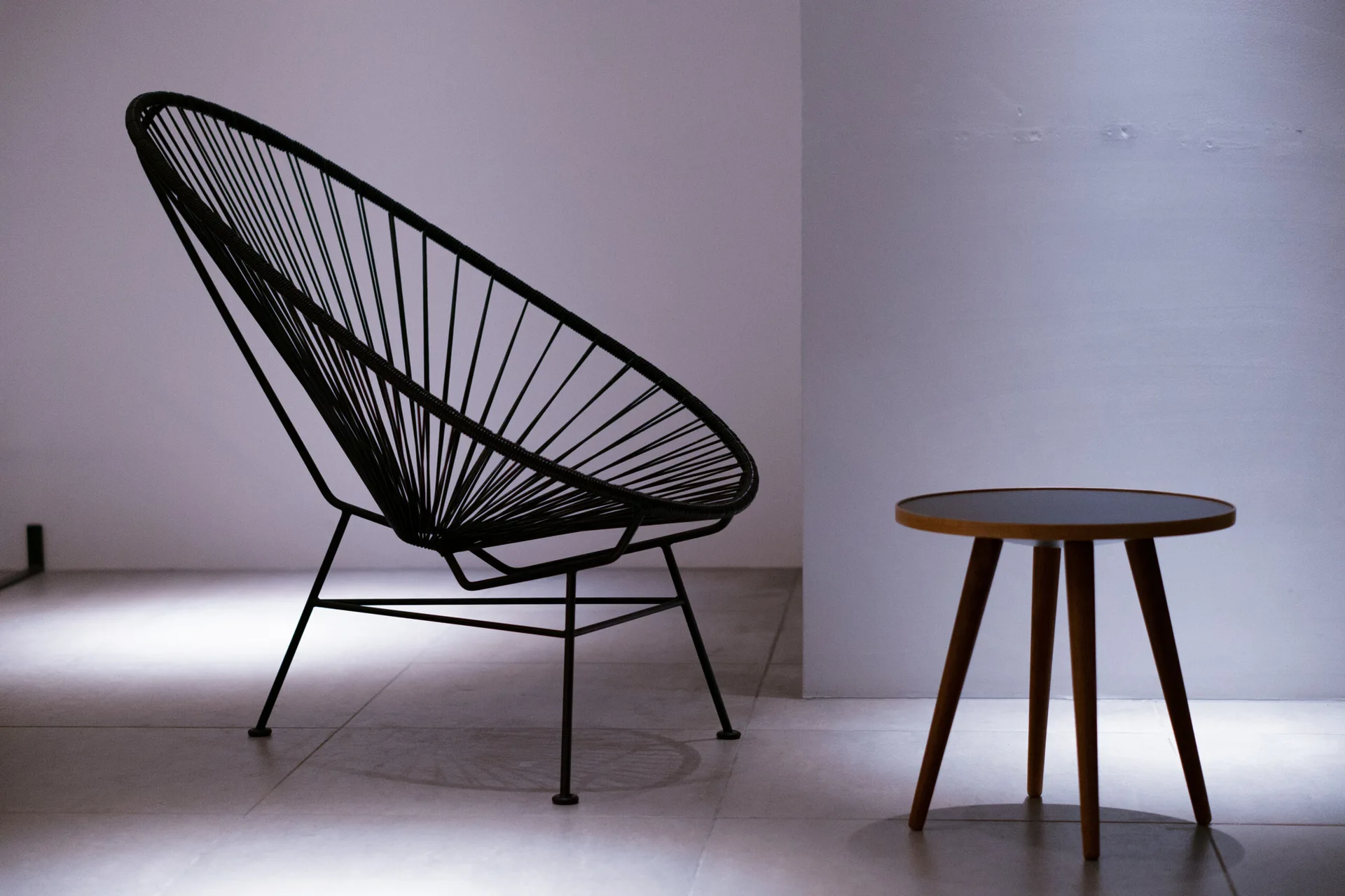 A black woven Acapulco chair and a round wooden side table stand on a grey tiled floor against a plain white wall.