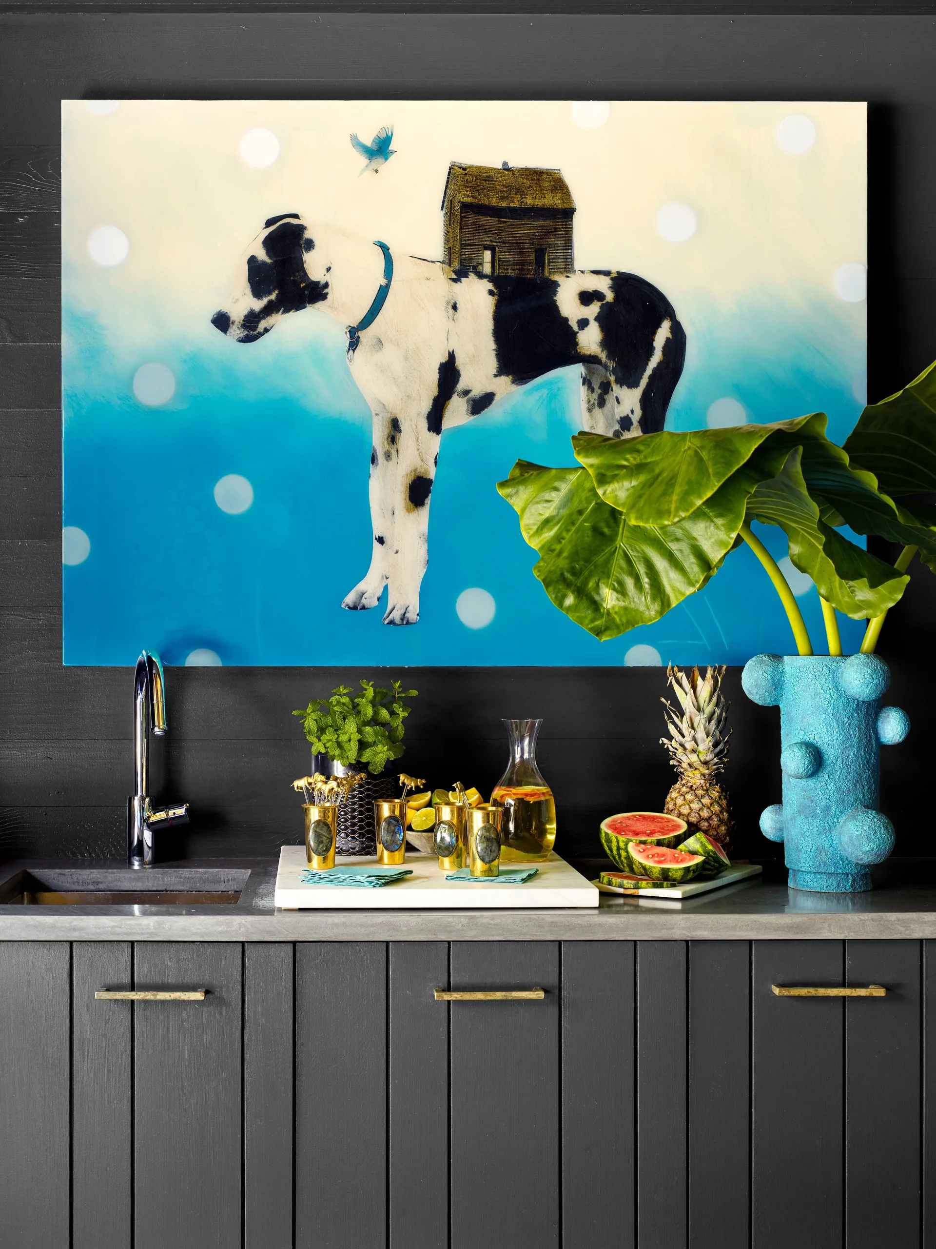 A dark grey wet bar features charcoal cabinetry, a stone countertop with a small sink, and a large blue painting of a dog hanging on a dark wood wall.