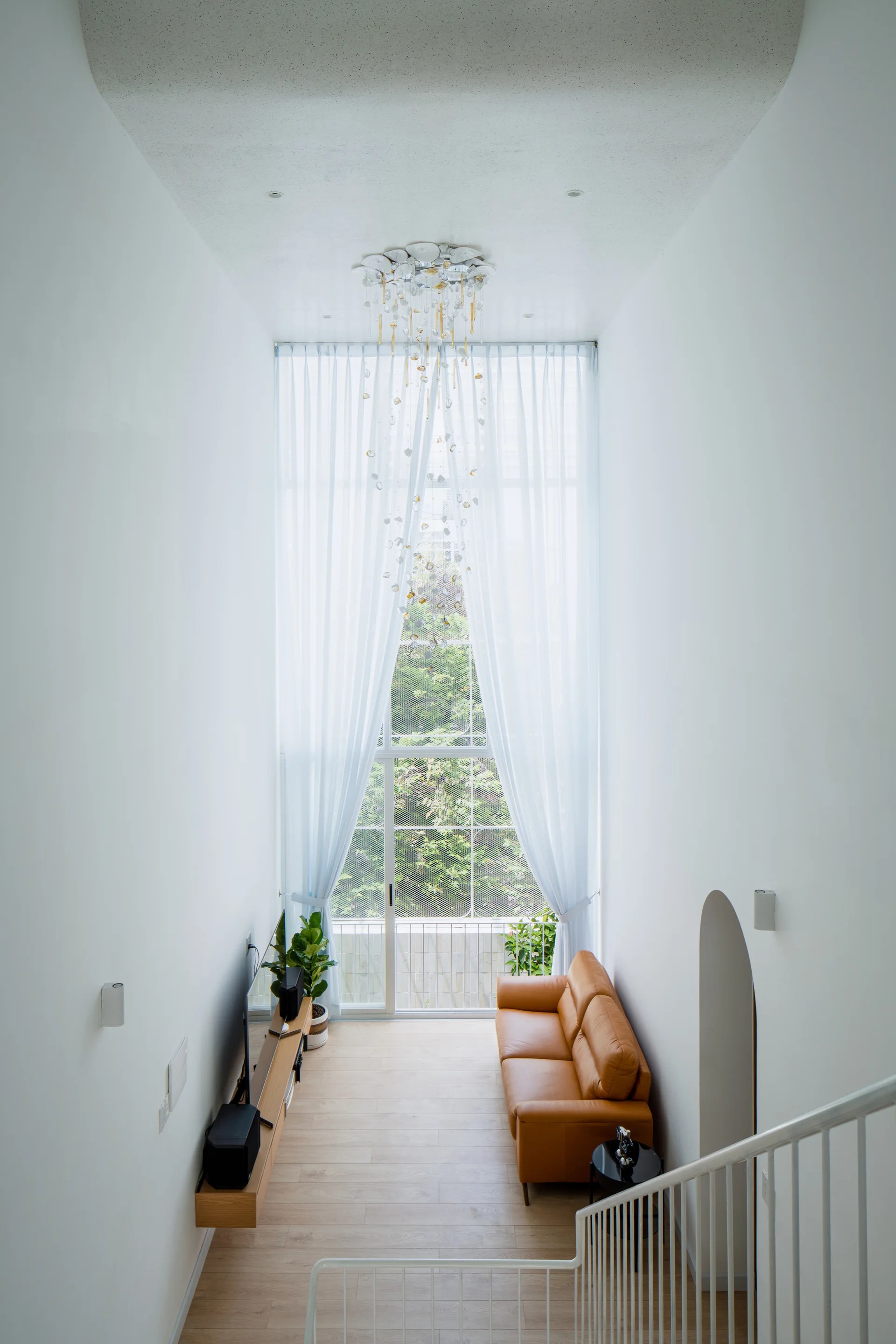 An elevated view of a narrow living room featuring a tan leather sofa, a floating wood console, and a tall window with sheer white curtains.