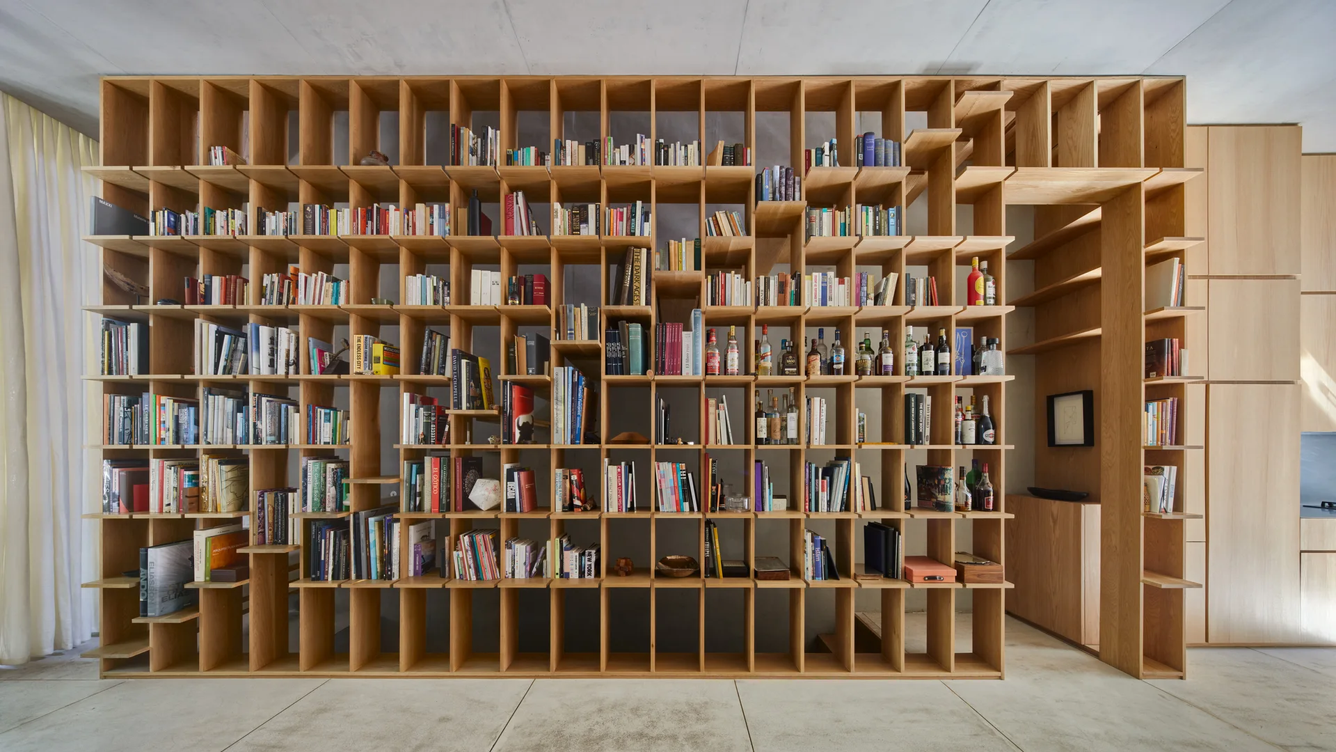 A large floor-to-ceiling wooden grid bookshelf filled with books and bottles sits against a concrete wall next to matching wood cabinets.