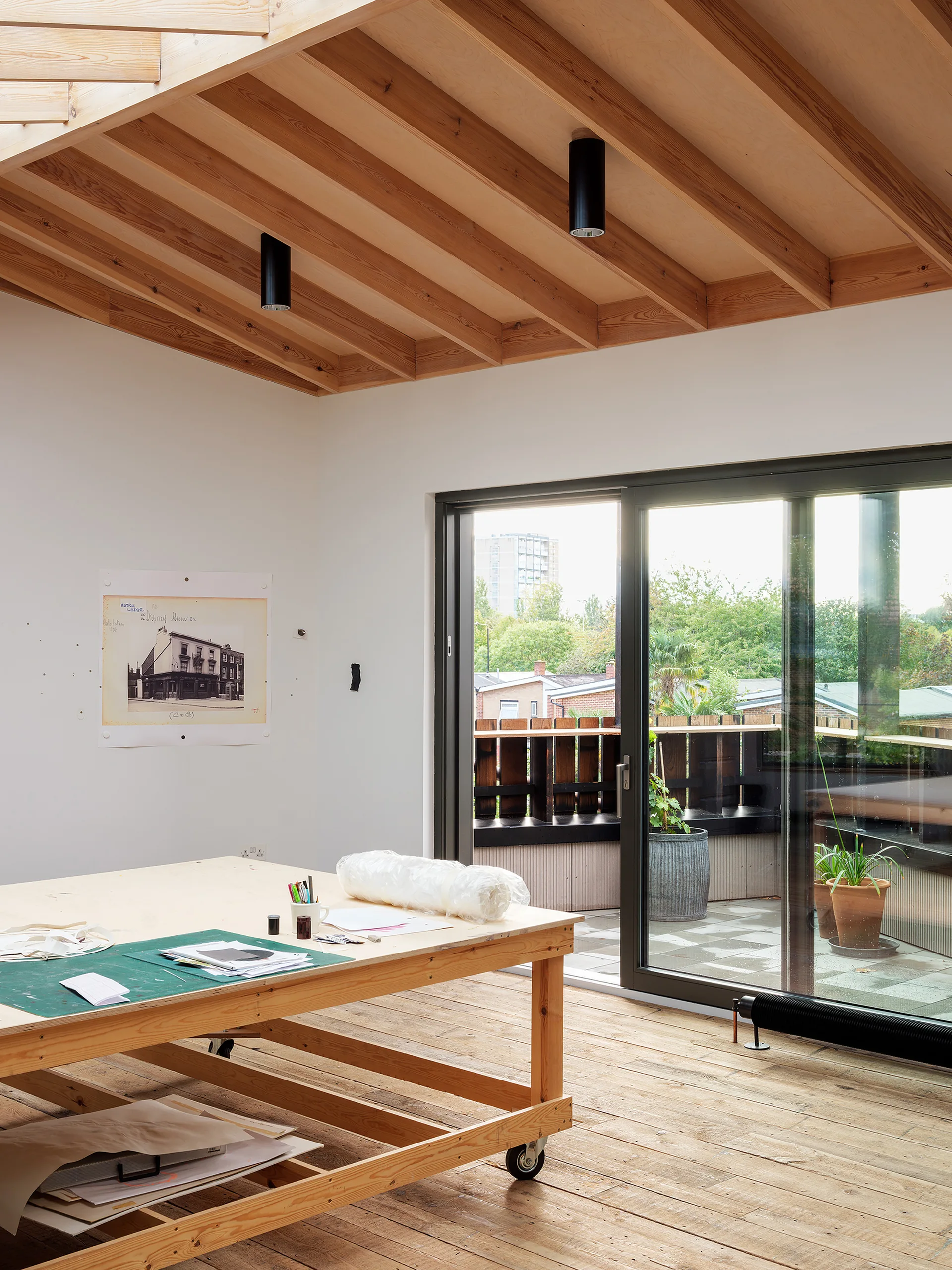 A bright studio workspace features a large wooden table on wheels under an exposed timber ceiling next to sliding glass doors that open to a balcony.