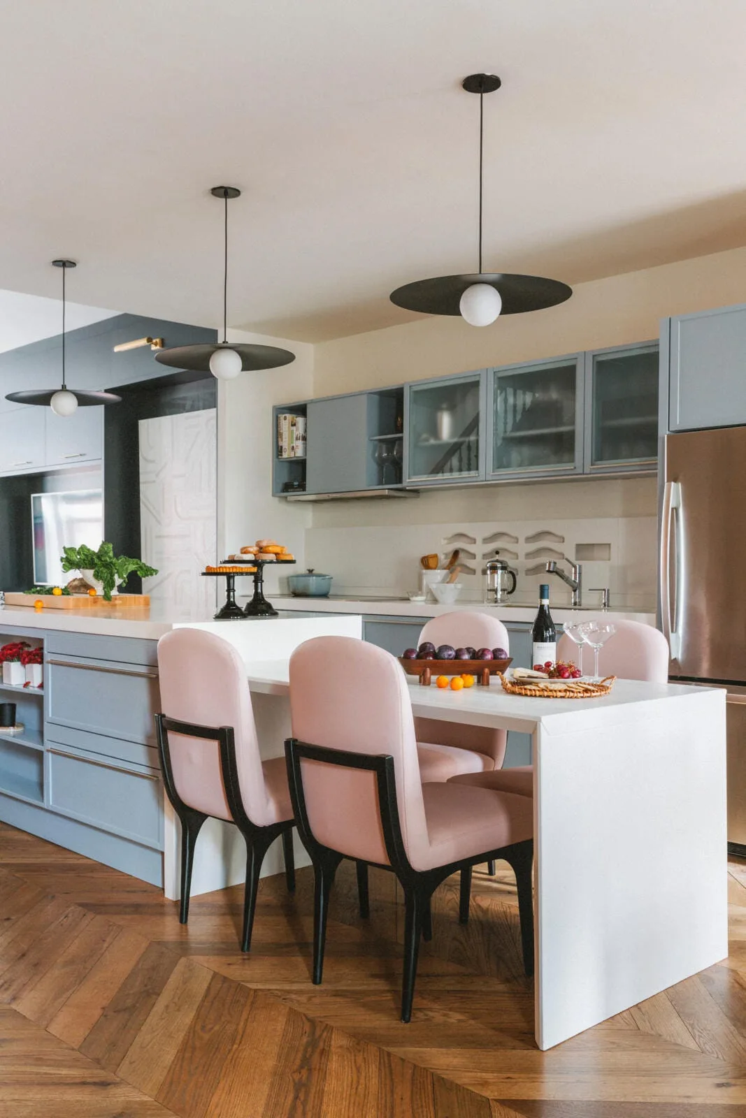 A modern kitchen features light blue cabinetry, a white island with pink upholstered dining chairs, black pendant lights, and herringbone hardwood floors.