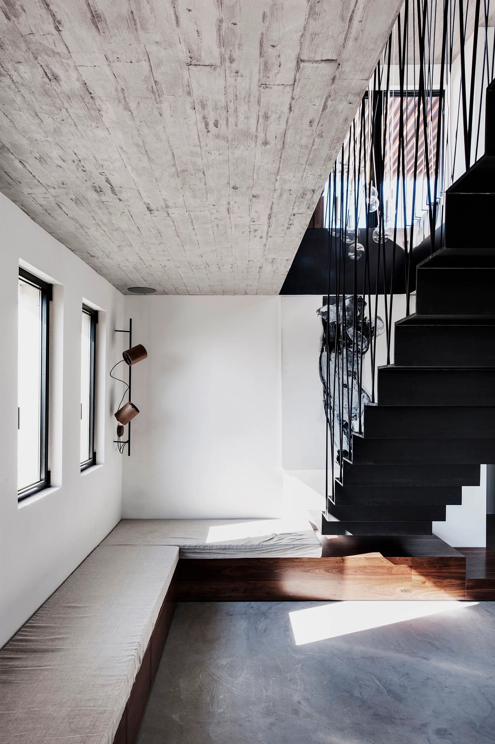 A modern interior entryway features a floating black metal staircase beside a low wood built-in corner bench under a board-formed concrete ceiling.