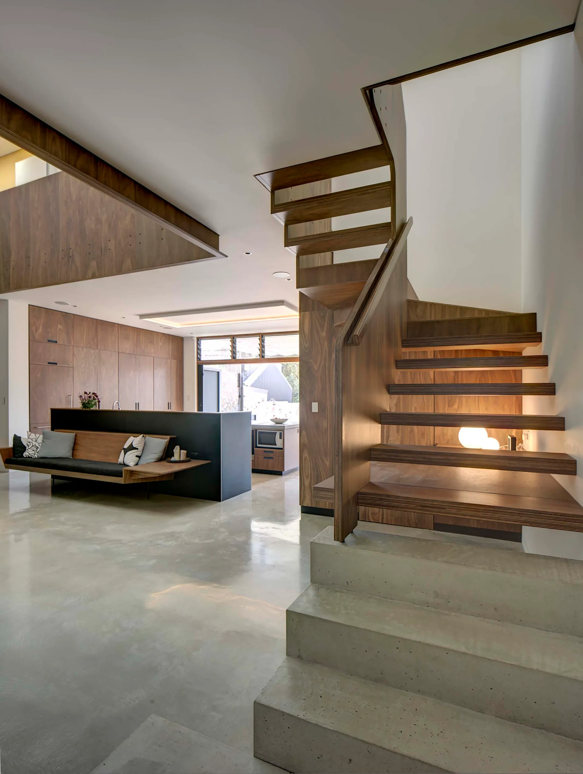 An open-plan room features a floating wooden staircase rising from concrete base steps, set against polished concrete floors and matching wood cabinetry.