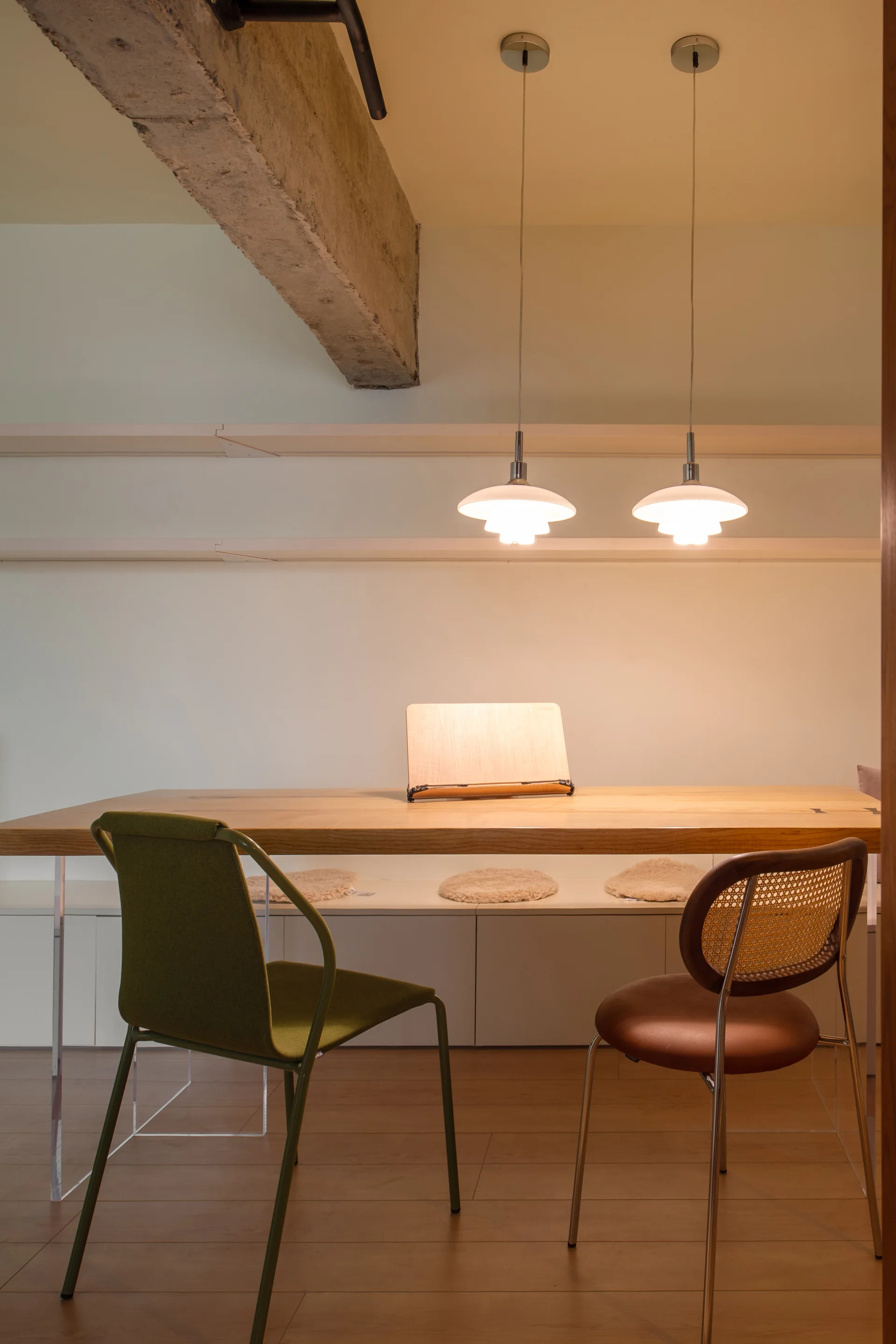 A wooden dining table with transparent acrylic legs sits below an exposed concrete beam and two glowing white pendant lights.