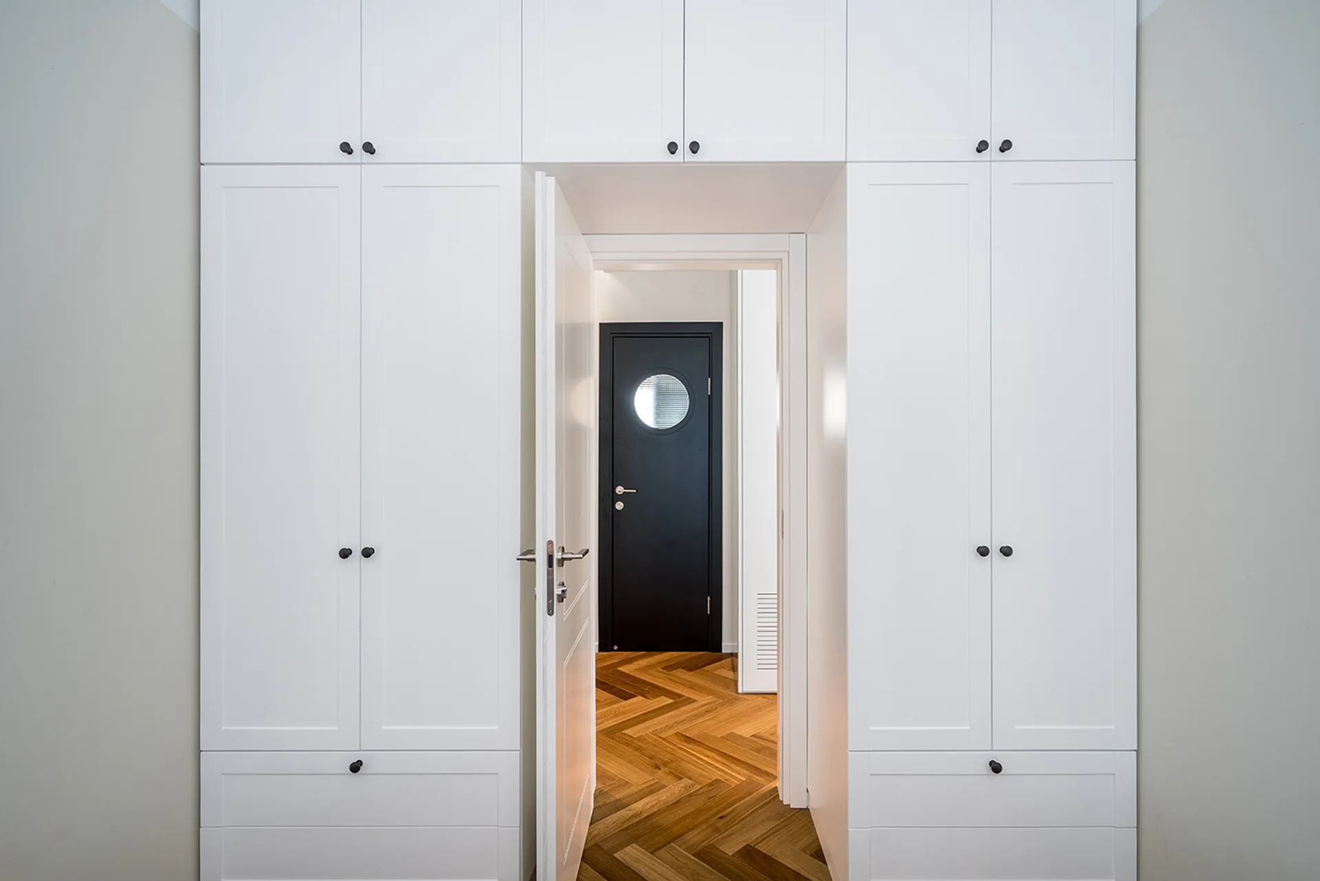 White floor-to-ceiling built-in cabinets frame a doorway looking into a hall with herringbone wood flooring and a dark door with a round window.