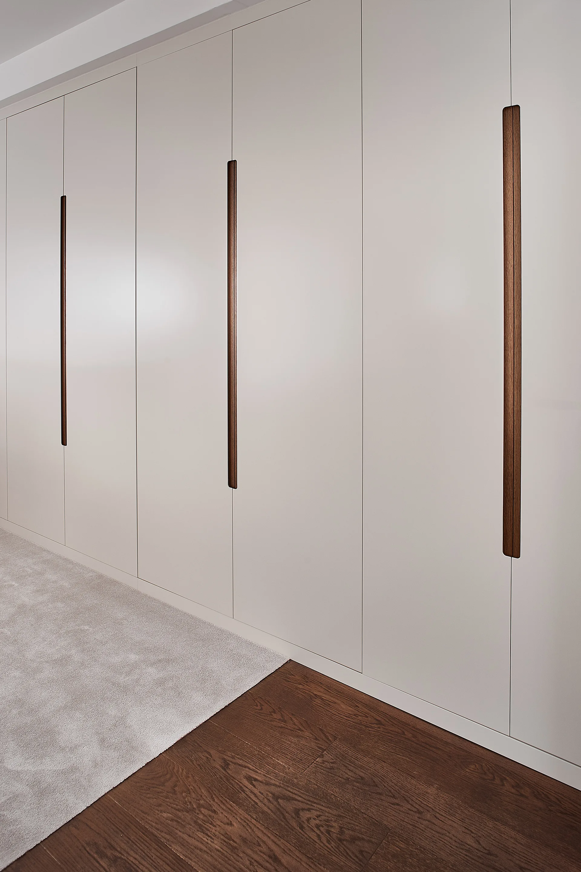 A wall of floor-to-ceiling cream cabinetry featuring inset vertical wood handles next to a wooden floor and grey rug.