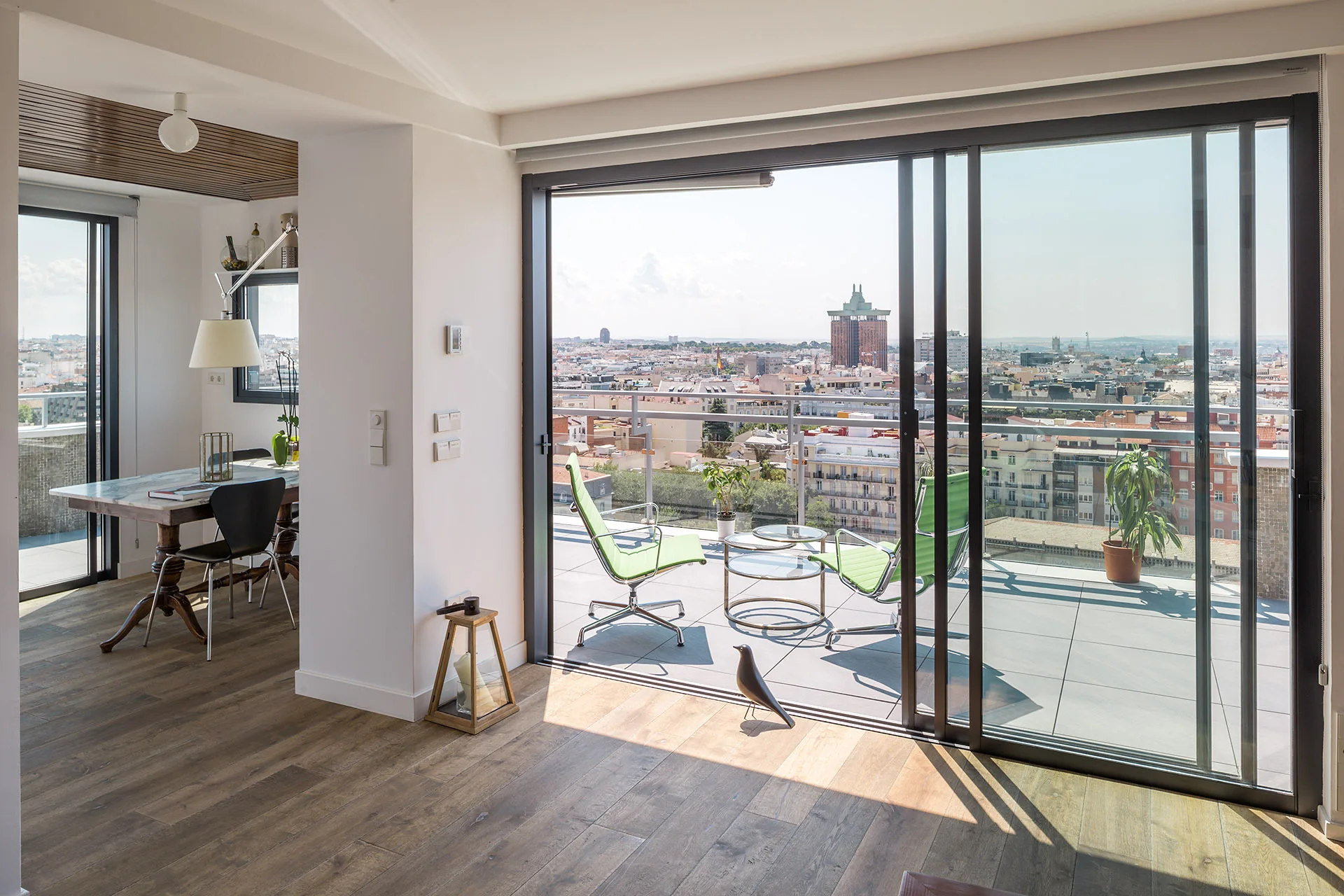 A modern dining area with wood flooring and a marble-top table opens through sliding glass doors to a sunny terrace with green chairs and a city view.