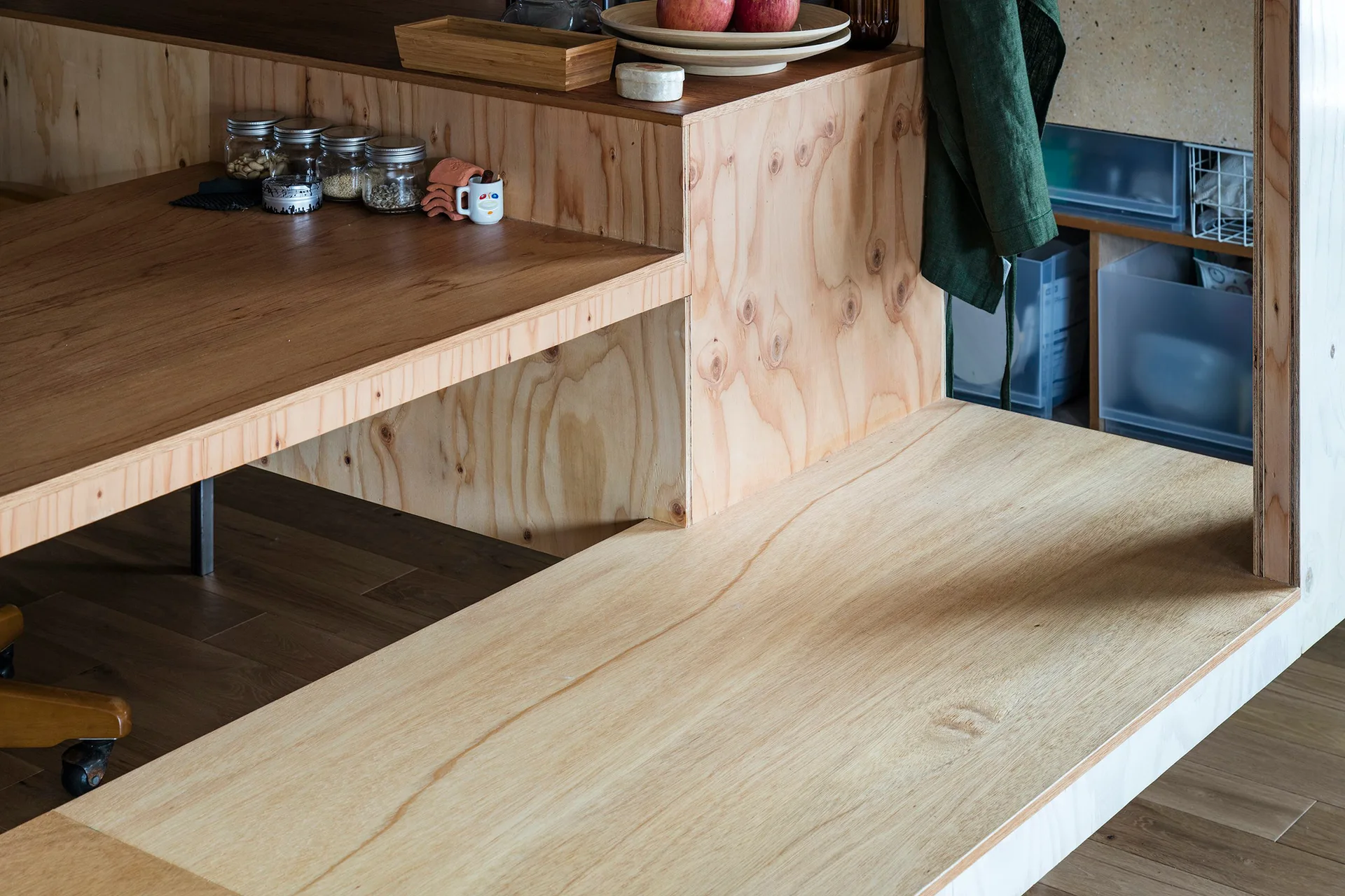 A close-up of a multi-level built-in plywood shelving unit and desk displaying glass jars, ceramic bowls, and hanging green linen.