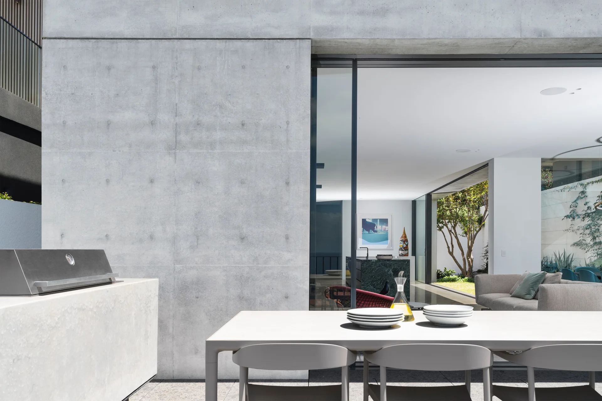 A modern outdoor patio features a light gray dining table and chairs set against an exposed concrete wall next to a large sliding glass door.
