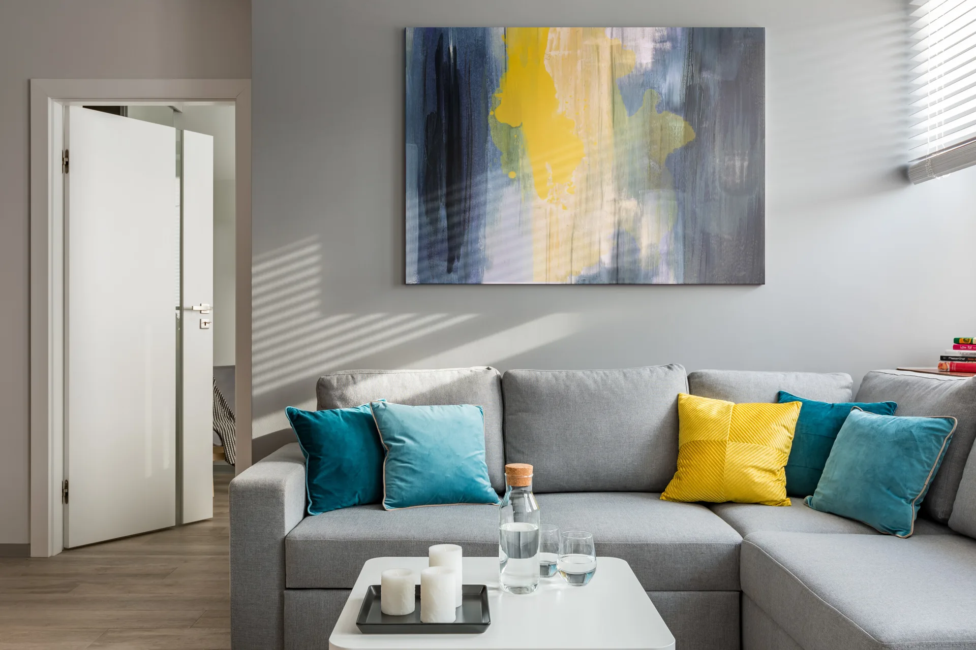 A modern gray sectional sofa is decorated with teal and yellow pillows beneath a large blue and yellow abstract painting.