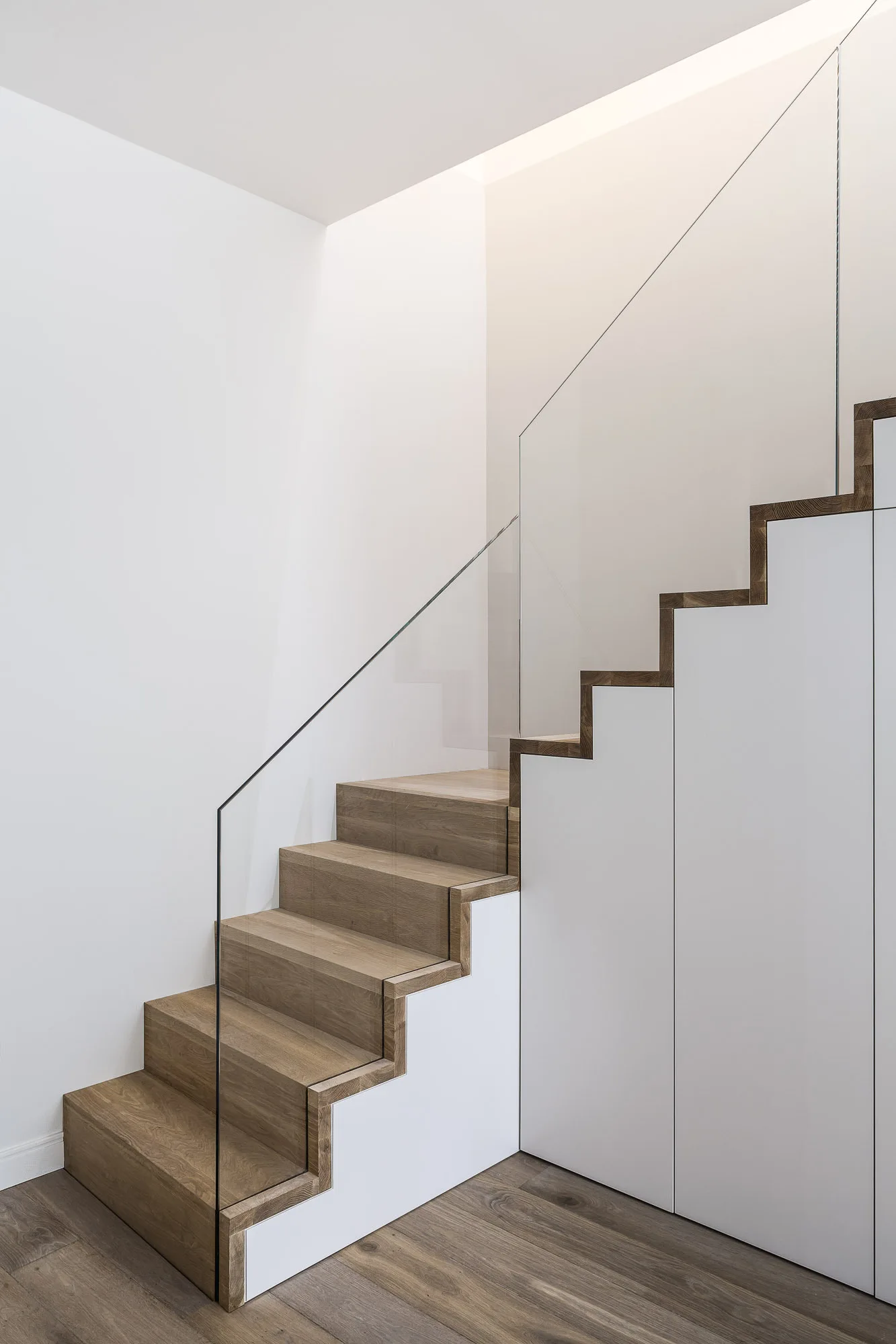 A modern wood staircase with a frameless glass railing and white storage cabinets integrated flush underneath the treads.