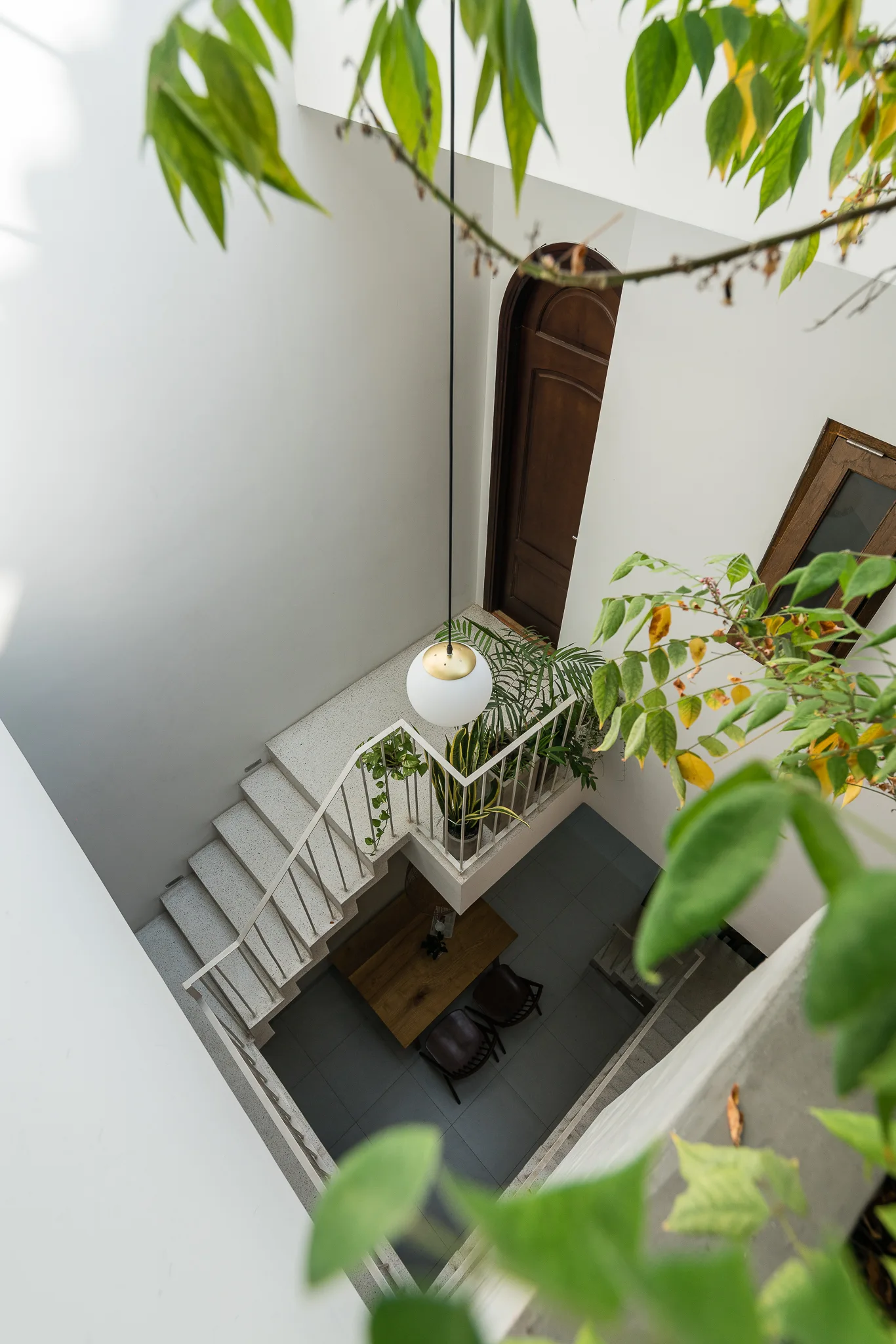 A high-angle view of a modern white staircase curving down to a dining area with a hanging globe pendant and potted green plants.