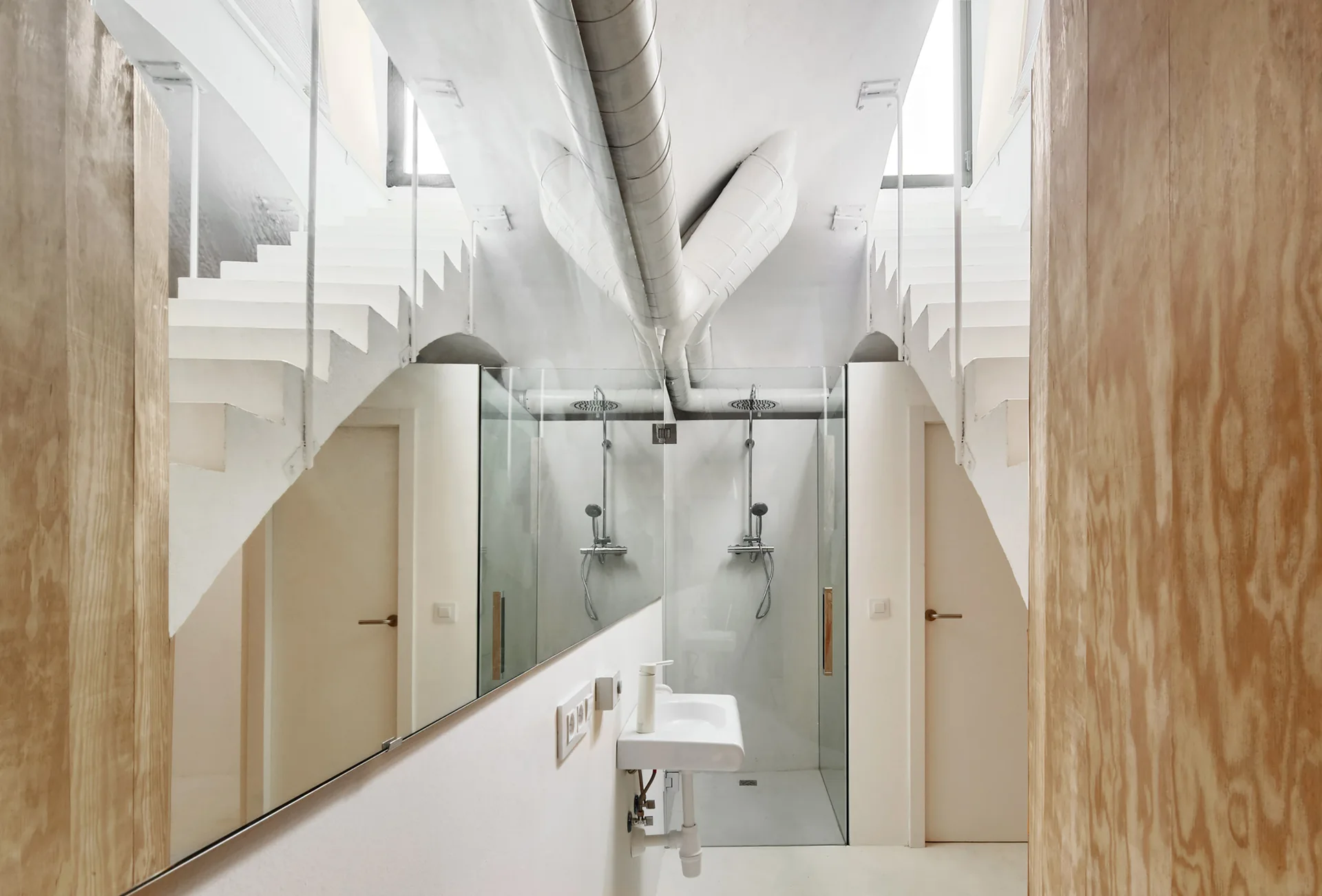 A minimalist white bathroom featuring a glass shower, a wall-mounted sink, a large mirror, plywood wall panels, and exposed silver ceiling ductwork.