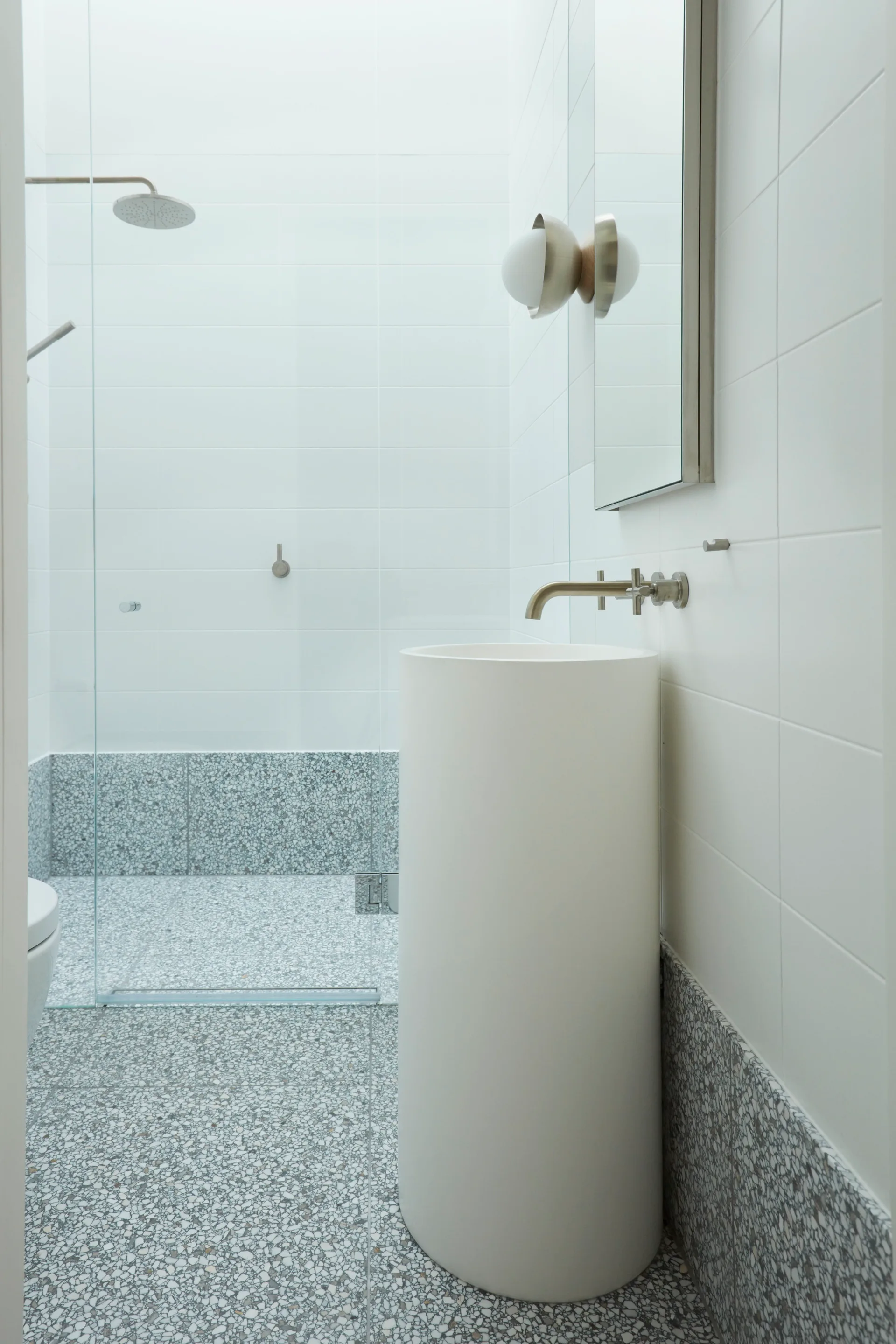 A minimalist bathroom features a white cylindrical pedestal sink, wall-mounted faucet, and grey speckled terrazzo flooring.