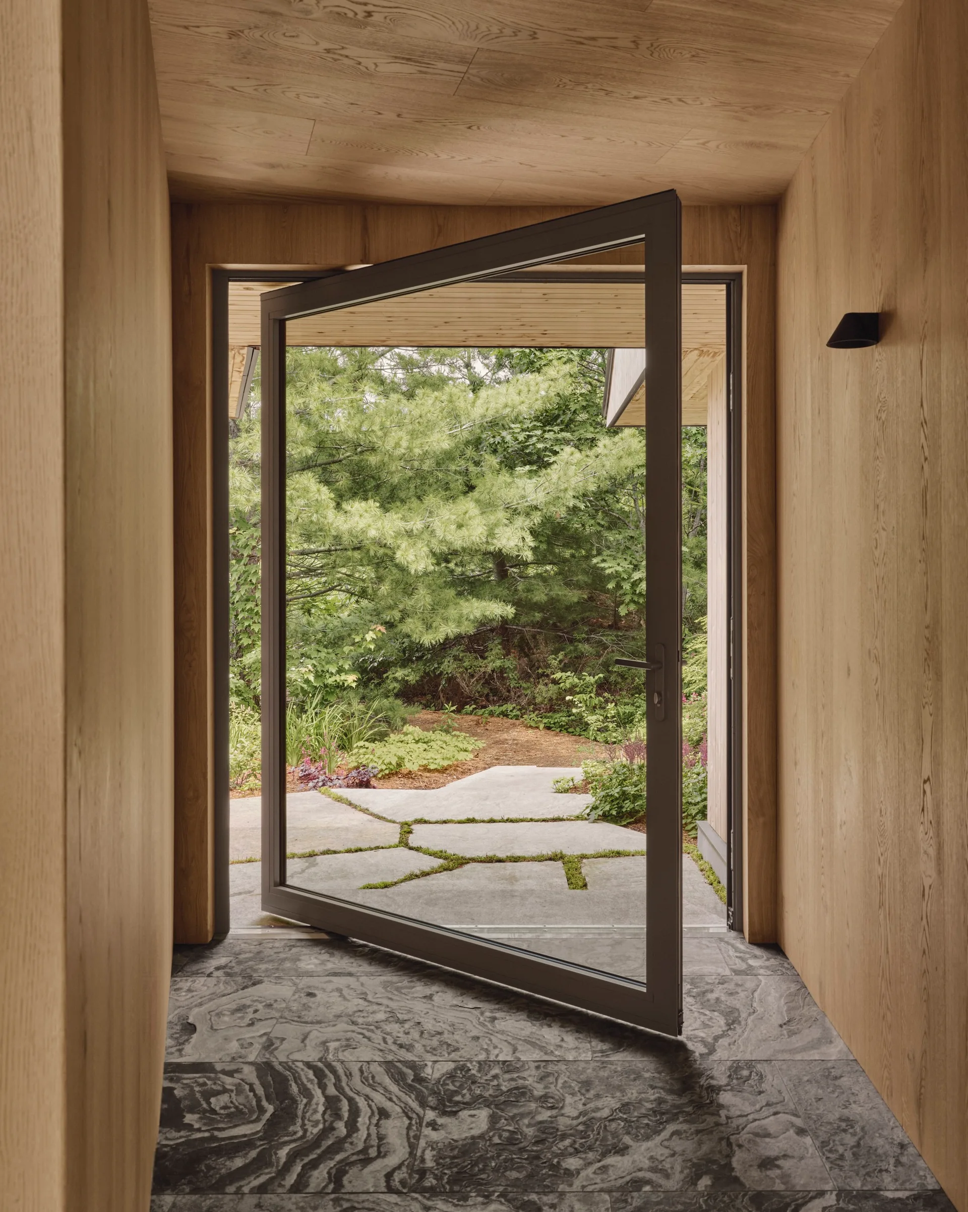 A wood-paneled entryway features grey veined stone flooring and a large glass pivot door opening to a flagstone garden path.
