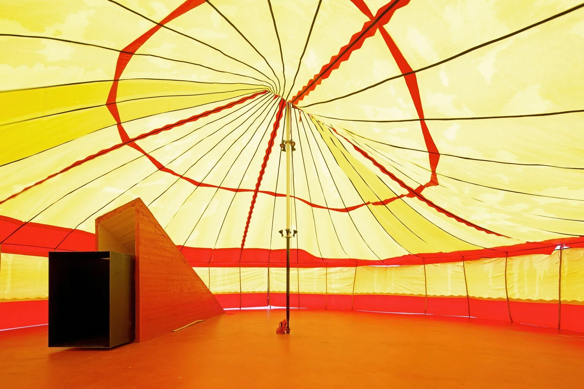 The interior of a large circular tent features a yellow and red fabric canopy, a central metal support pole, and a bright orange floor.