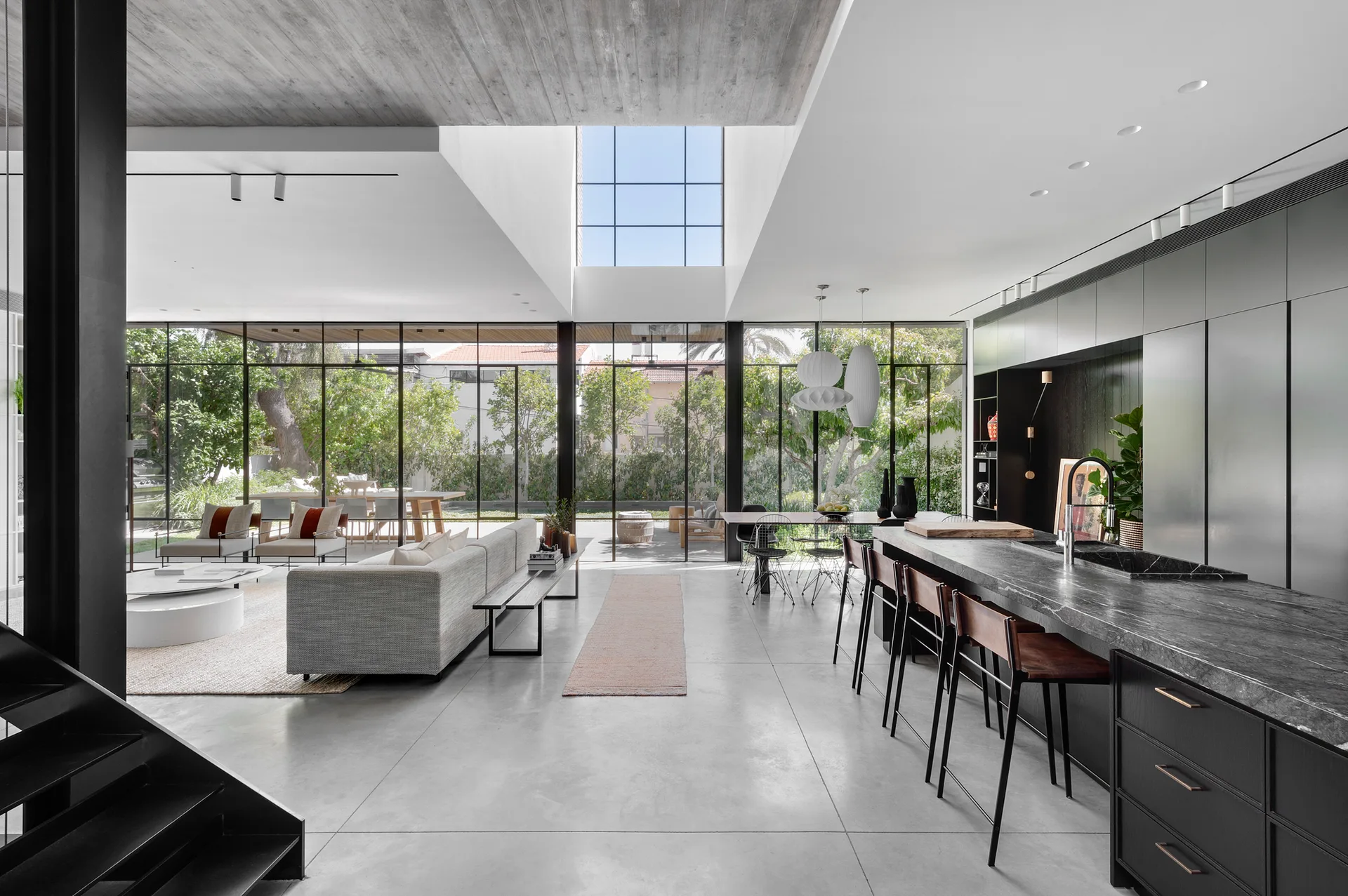 A modern open-plan kitchen and living space featuring a dark marble island, gray concrete floors, and floor-to-ceiling glass walls overlooking a garden.