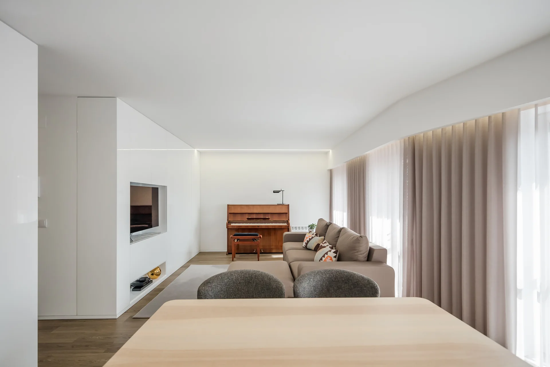 A contemporary living room featuring a beige sectional sofa, a wooden piano, and custom floor-to-ceiling white media cabinetry.
