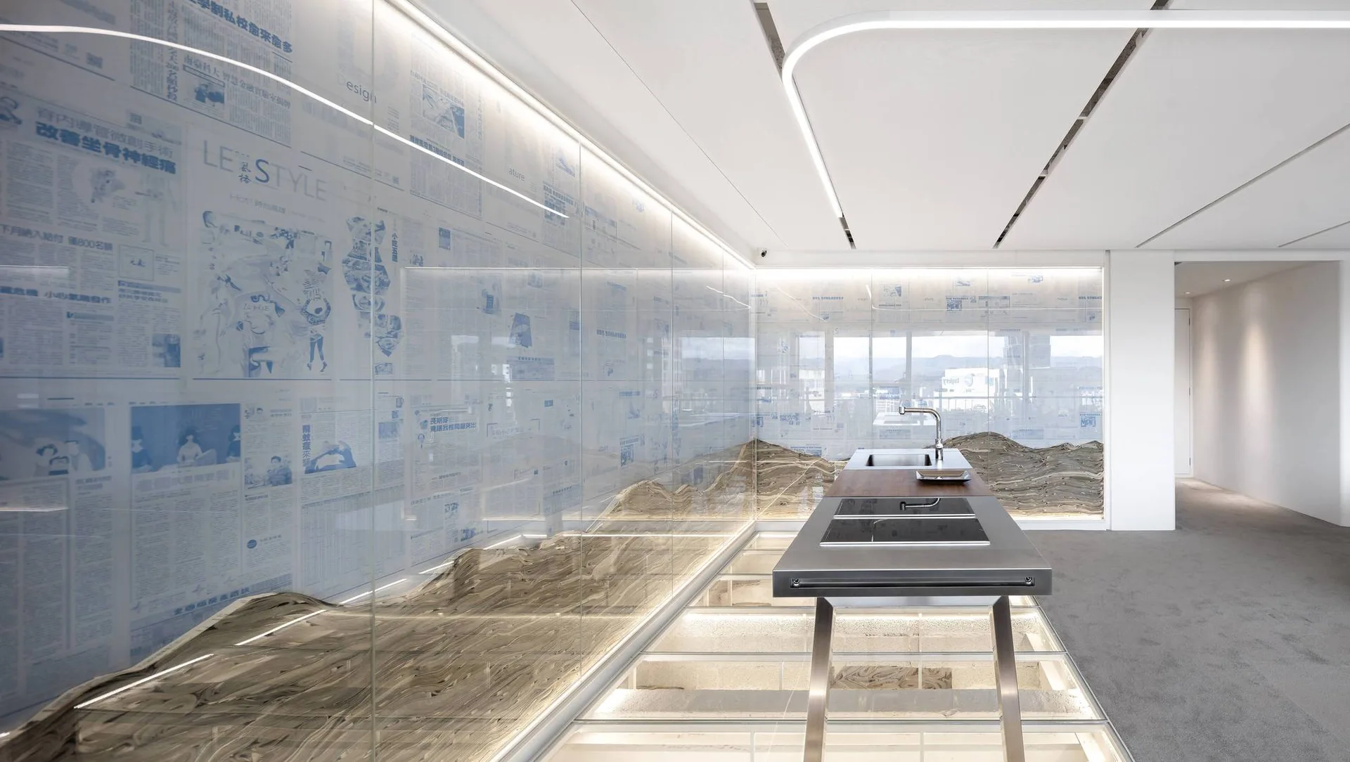 A minimalist kitchen features a stainless steel island next to an illuminated glass floor and a glass partition wall printed with newspaper articles.