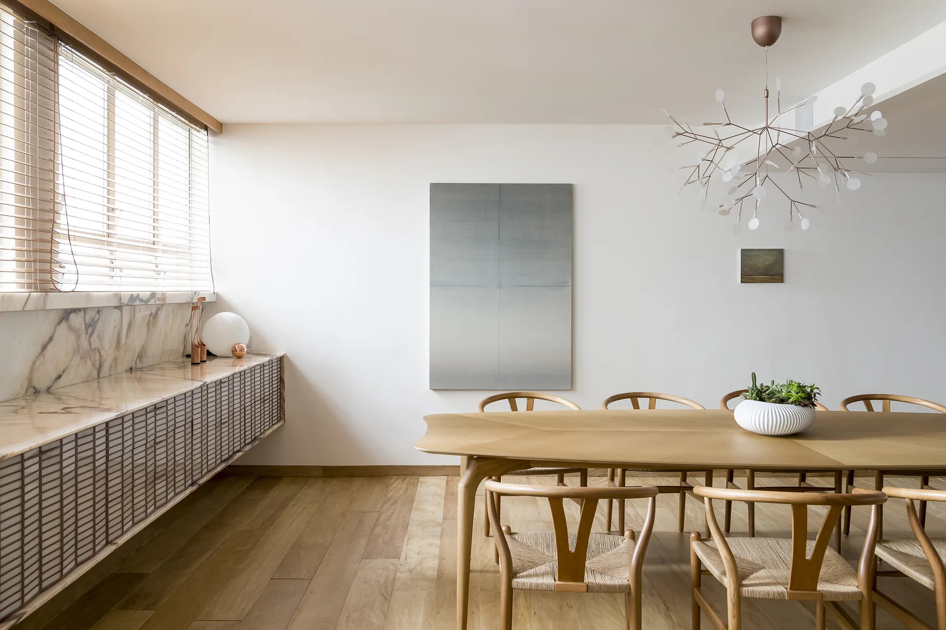 A minimalist dining room featuring a light oak table surrounded by woven-seat wishbone chairs beneath a branching white chandelier.