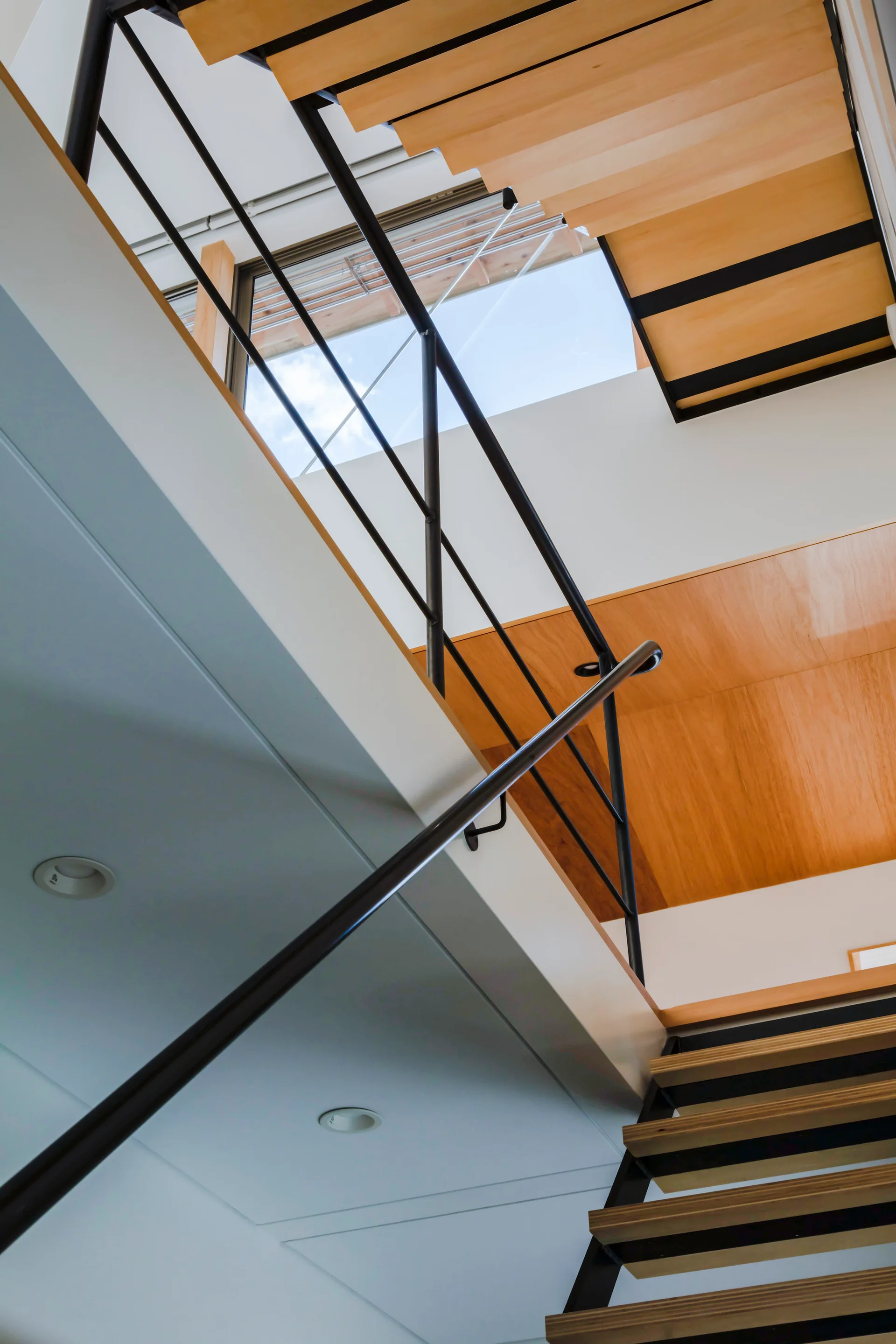 Looking up a modern staircase featuring light wood treads, black metal railings, a wood-paneled ceiling section, and a high window showing the sky.