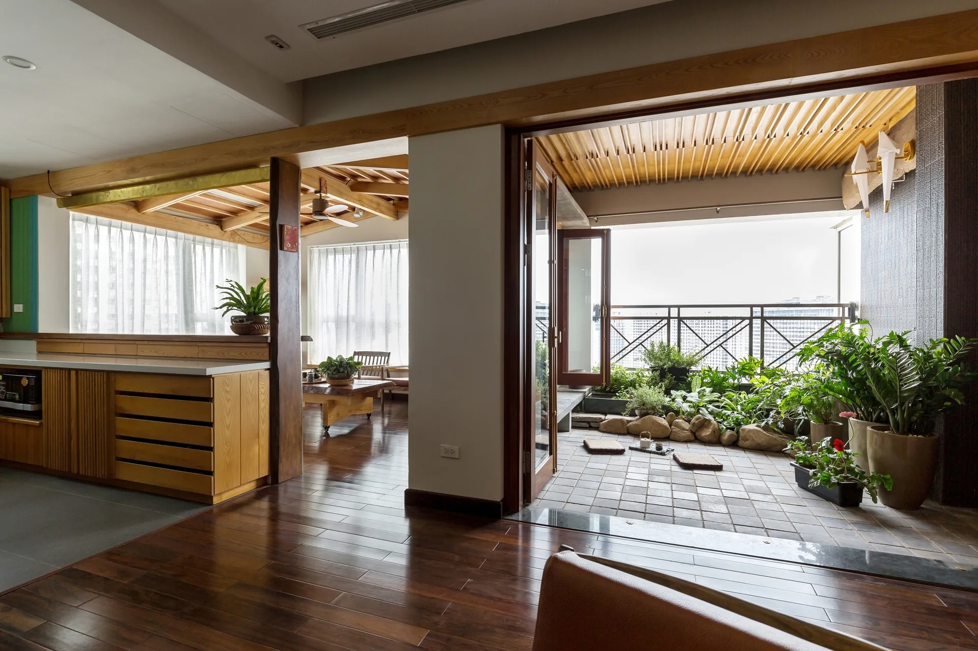 A modern apartment interior with dark wood floors opening onto a stone-tiled terrace with lush green potted plants and floor cushions.